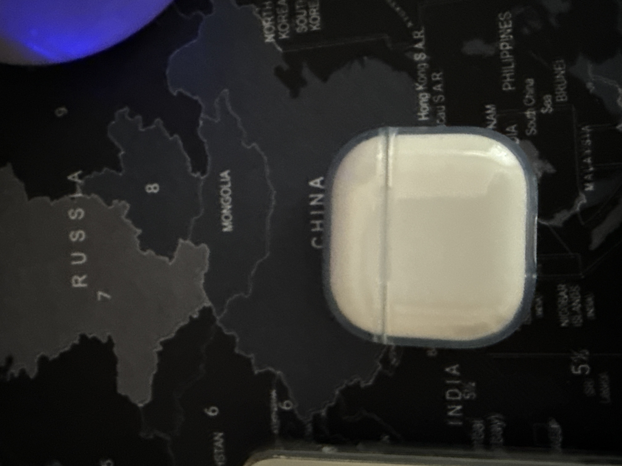 The image shows a white, square-shaped object with rounded corners, possibly a case for electronic devices, resting on a dark map. The map features the countries of Russia, Mongolia, China, India, and parts of Southeast Asia including the Philippines and Malaysia, along with surrounding bodies of water. Text labels identifying the countries and regions are visible on the map. A partially visible blue light source is present in the upper left corner of the frame. There is no information about the city of Lovech, Bulgaria, or any other location.