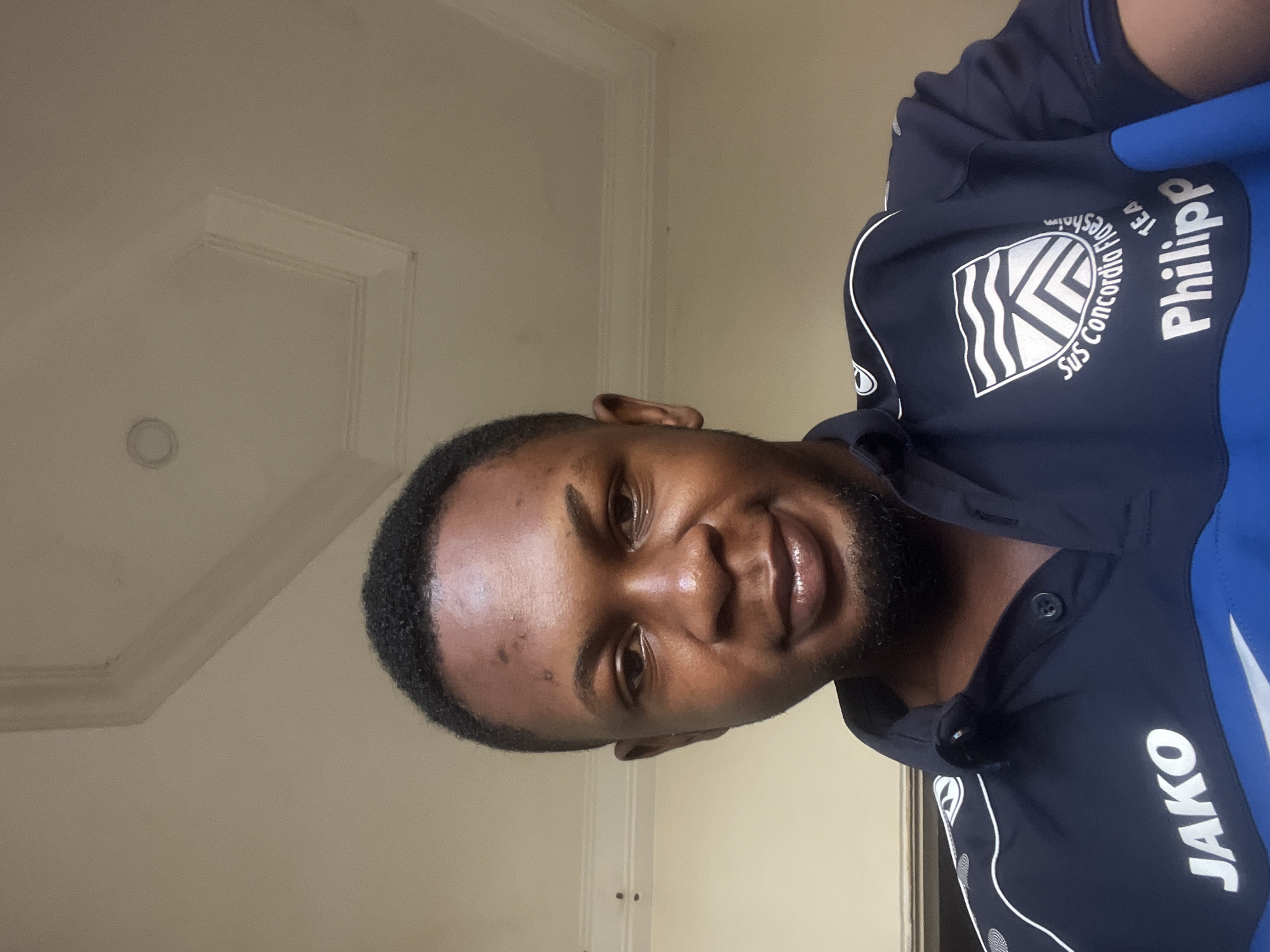 This is a selfie of a man indoors, facing the camera. He has dark skin, short black hair, and a short beard. He is wearing a dark blue polo shirt with white trim and logos. On the left side of the shirt, the word "JAKO" is printed in large white letters. On the right side, there is a shield-shaped logo with wavy lines and an emblem, and below it, the name "Philipp" is printed in white letters. The man is smiling slightly. The background appears to be a room with a beige wall and a decorative ceiling with recessed lighting. There is no specific indication of the location being Jalingo, Nigeria within the image itself.