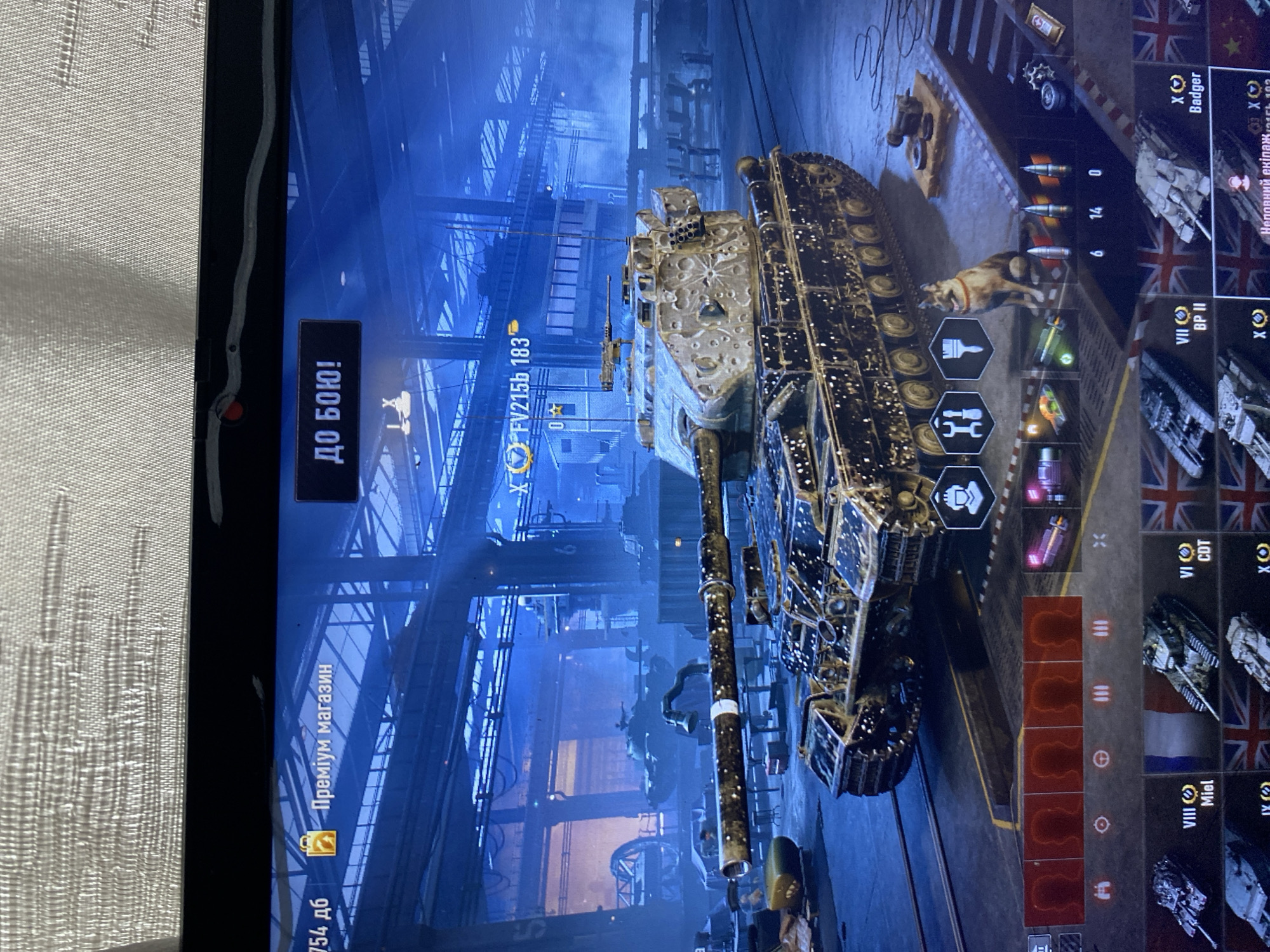 This is a screenshot of a video game, likely World of Tanks, displayed on a laptop. The game features a heavily decorated tank in the foreground, within what appears to be a virtual garage or workshop environment. The background shows a dimly lit industrial setting with blue lighting, possibly depicting a factory or hangar. There are other tanks and machinery visible in the background.
The user interface elements of the game are visible, including buttons labeled "ПРЕМІУМ МАГАЗИН" (Premium Shop) and "ДО БОЮ!" (To Battle!). There is also information about a specific tank, "FV215b 183". Below the main tank, there are icons representing crew members and ammunition. Further down, a selection of other tanks is displayed. A small dog is sitting on the ground near the main tank.
There is no indication in the image that this scene is located in the city of Szczecin, Poland. The image depicts a virtual environment within a video game.