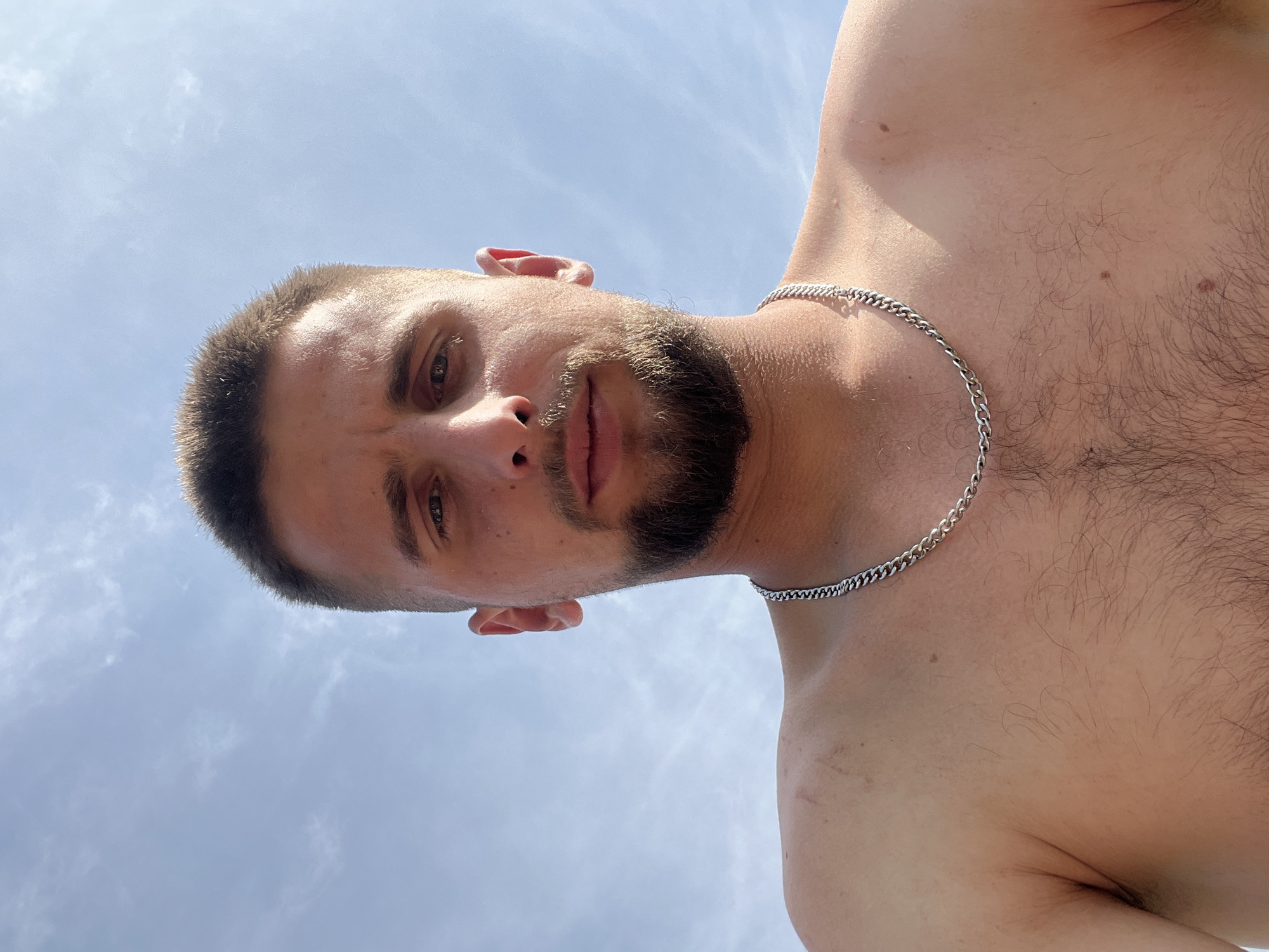 An upward-angle shot depicts a shirtless adult male looking directly at the viewer against a bright blue sky with faint cirrus clouds. The subject has short, dark brown hair, light eyes, and a trimmed beard. A silver-colored curb chain necklace is worn around his neck. His chest displays moderate dark brown hair, and his skin exhibits multiple moles and faint reddish marks on the shoulders. The scene is outdoors in Kapustino, Russia.