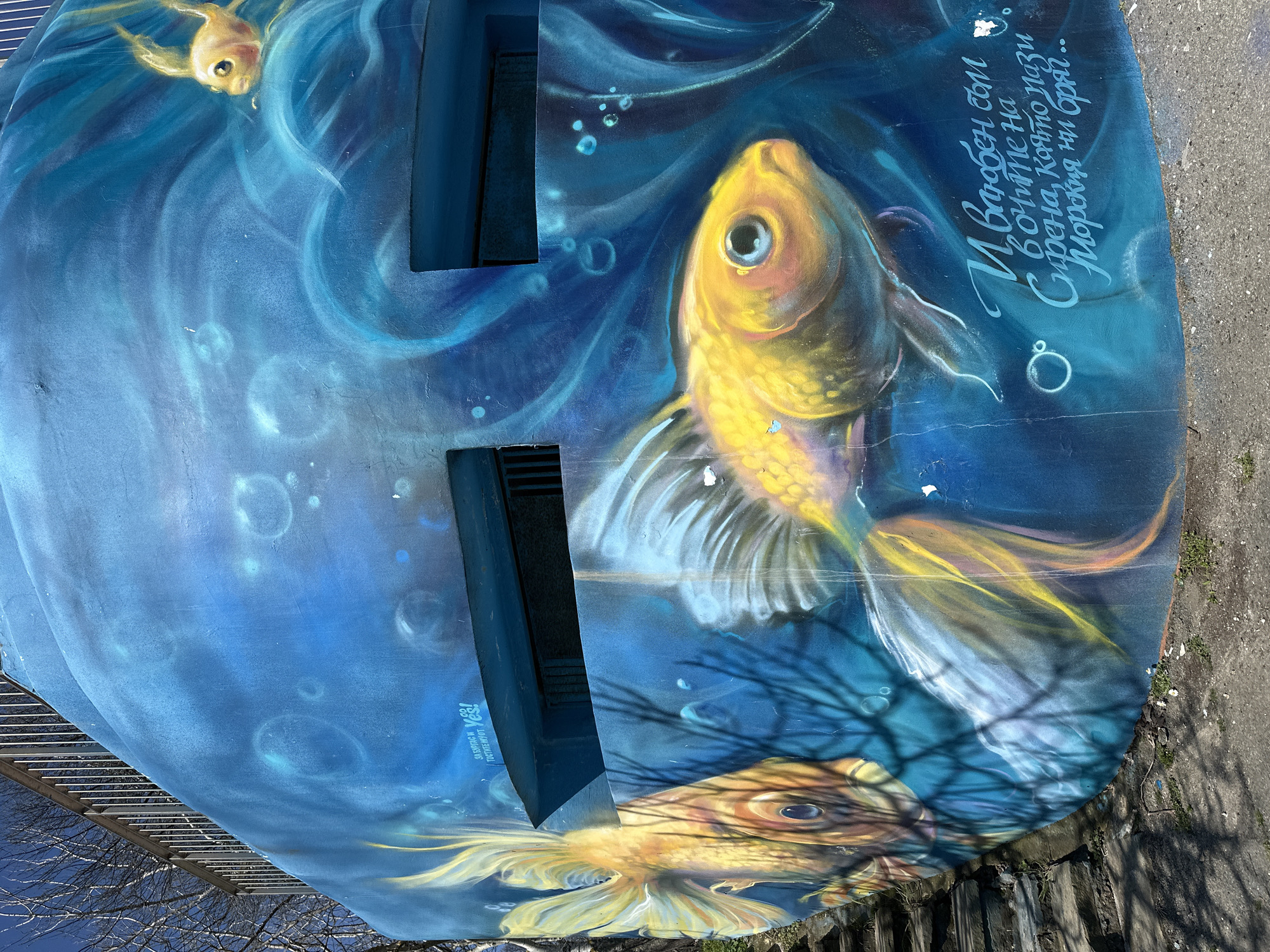 The image shows a section of painted artwork on a structure with air vents. The artwork is predominantly blue and depicts underwater scenes with three golden fish. Bubbles and stylized water currents are also visible. Text in Cyrillic script is located on the right side of the artwork. The text is a verse about the city of Burgas and its sea coast. The text includes the name “Sirena” (mermaid).
The bottom edge of the painted structure is against a gray walkway with fence and some vegetation. The structure has the words "ZA BURGAS I 100" and "Yes!"