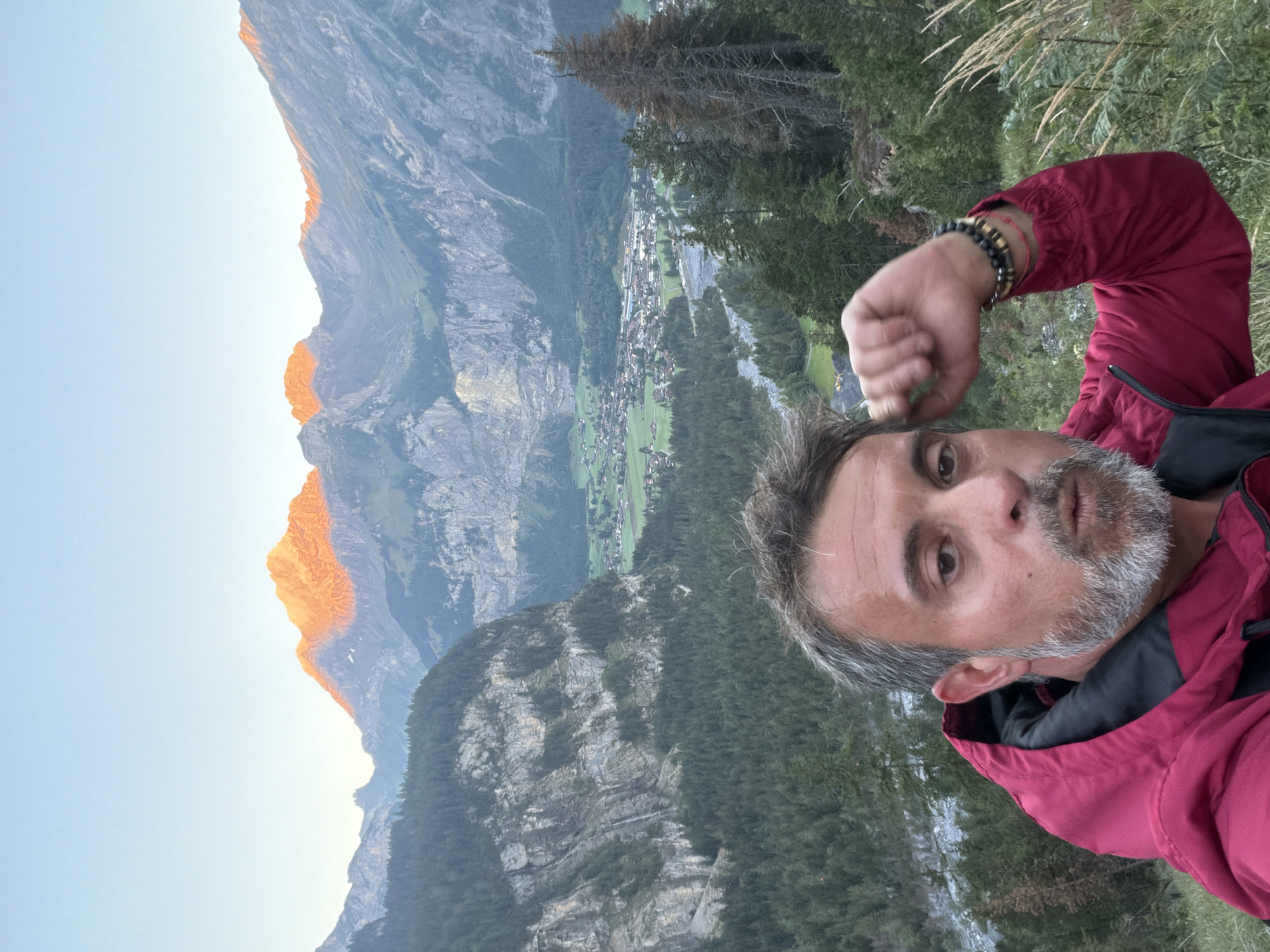 The image is a selfie taken by a man with graying hair and a beard, wearing a red jacket. He is in the foreground, looking at the camera with his mouth slightly open. His right arm is raised, with his hand near his head. Behind him, a valley unfolds, with a river flowing through it and a small village nestled amidst green fields. Towering mountains form the backdrop, with the peaks illuminated by the golden light of sunset. The environment is mountainous and rural, possibly in the Swiss Alps, given the mention of Kandersteg. The overall impression is one of a traveler capturing a scenic moment during a hike.