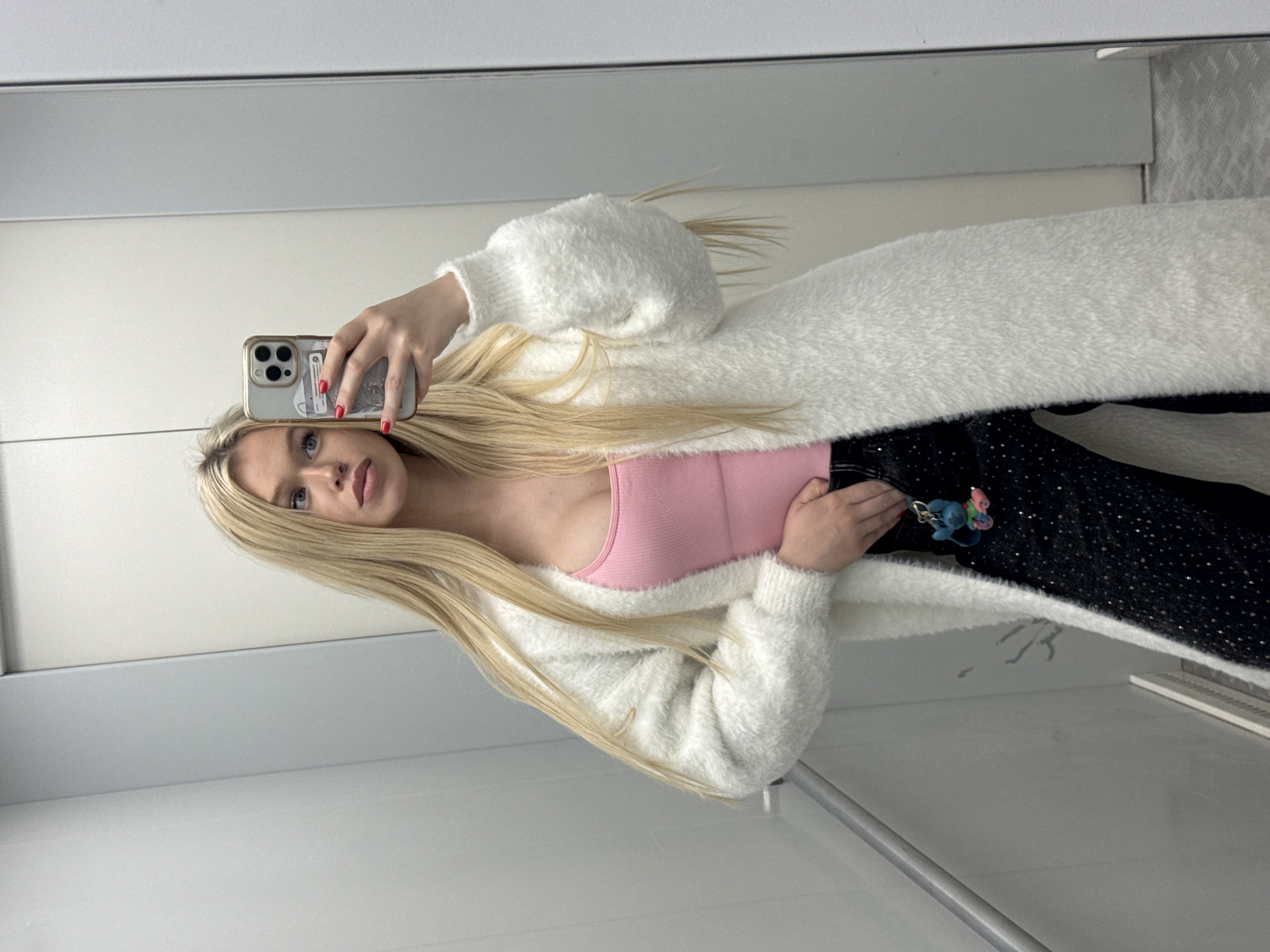 A young woman with long blonde hair is taking a mirror selfie in an elevator. She is wearing a pink tank top, a white fluffy cardigan, and black pants adorned with silver rhinestones. A keychain with a blue cartoon character is attached to her pants. The elevator appears to be modern and clean, with metallic walls and a tiled floor. The lighting is bright and even, typical of an indoor space. The location of Saint Petersburg, Russia, is not discernible from the visual information present in the image.