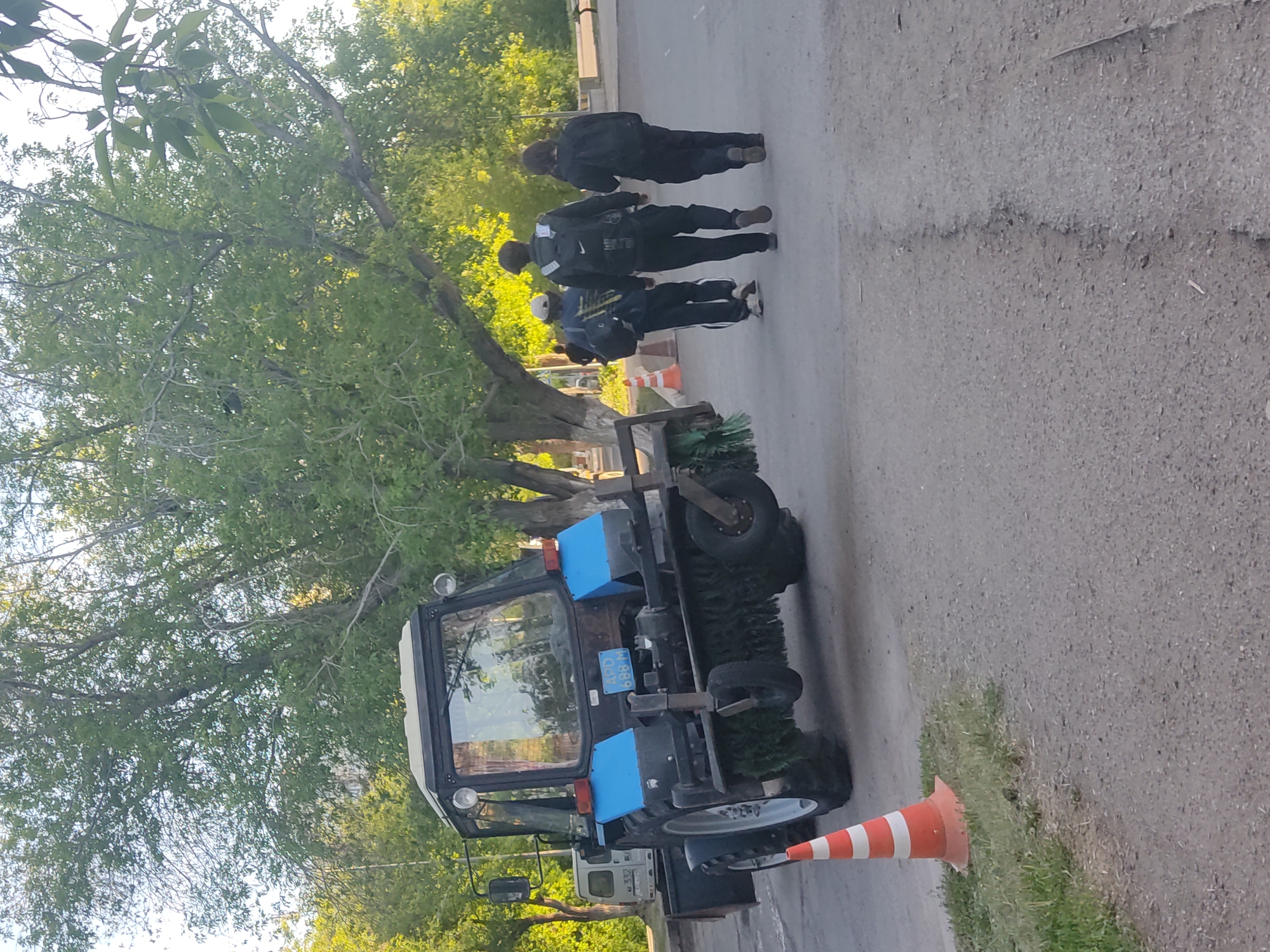 The image shows a street cleaning tractor parked on the side of a paved road in an urban or suburban area. The tractor has a blue and black color scheme and is equipped with rotating brushes for sweeping debris. A traffic cone stands next to the tractor. In the background, several people are walking away from the camera on the right side of the road. Trees line the side of the road. A white panel van is visible in the background. The location is in the city of Unknown, Unknown.