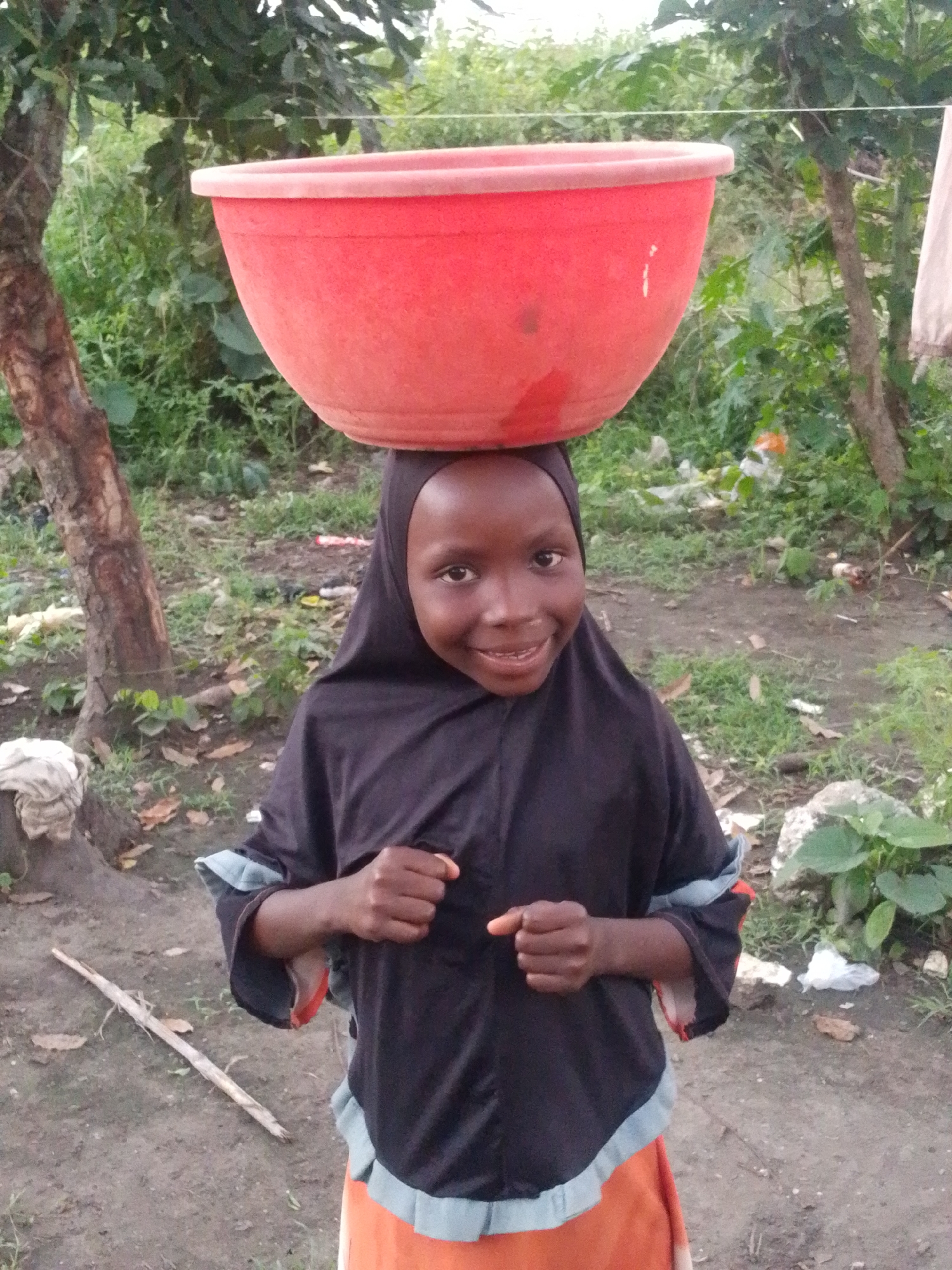 A young girl wearing a black hijab and an orange skirt stands outdoors in what appears to be the city of Otulu, Nigeria. She balances a large, red bowl on her head. The girl has a happy expression, with her mouth open in a smile, revealing her teeth. Her arms are held at her chest, with her hands clenched into fists. She is standing on a dirt path, with trees and greenery in the background. The ground is littered with leaves and debris. There is a thin white line stretching across the image behind the girl, possibly a clothesline. The lighting suggests it is daytime, possibly overcast.