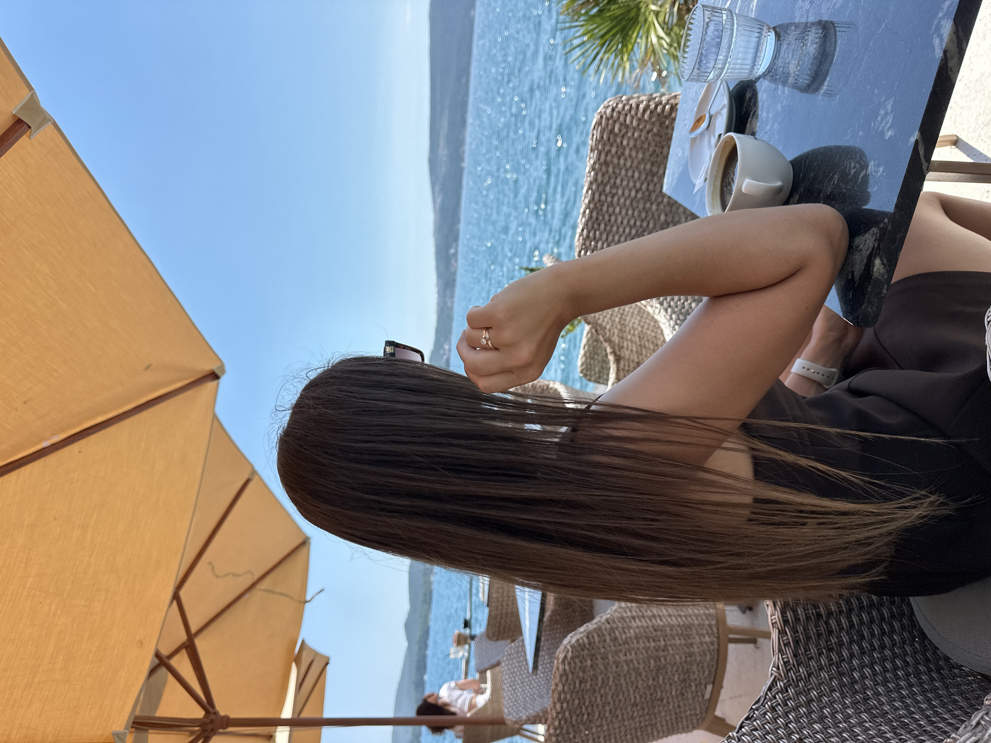 A woman with long, dark hair is seated at a table outdoors, facing away from the camera, with her profile visible as she looks out at a body of water. She is wearing sunglasses and a dark dress. On the table in front of her are a cup of coffee, a small plate with a spoon, and a glass of water. The scene appears to be a cafe or restaurant with tables and chairs, under an orange umbrella, overlooking a bay with mountains in the distance. The location is identified as Donja Lastva, Montenegro.
