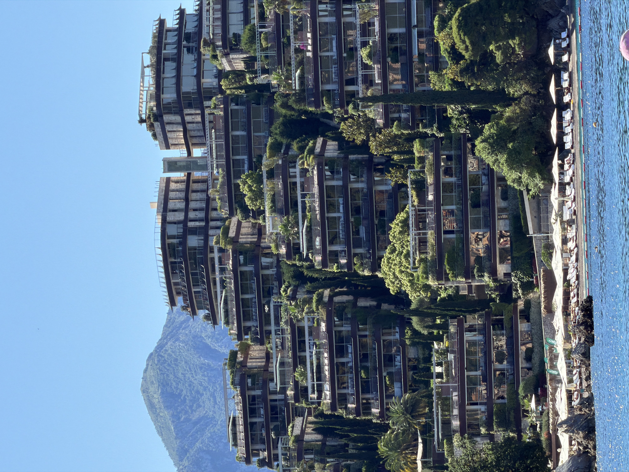 The image shows a high-rise building complex in Budva, Montenegro. The complex is multi-story with multiple levels of terraces adorned with greenery, including various trees and plants. The architecture features a modern style, with visible glass windows and steel or dark-colored frames. A mountain covered in dense forest is visible in the background, partially obscured by the building. In the foreground, there is a beach area with rows of sun umbrellas and loungers next to water that has a light blue tint. The sky is cloudless, which indicates bright and sunny conditions.