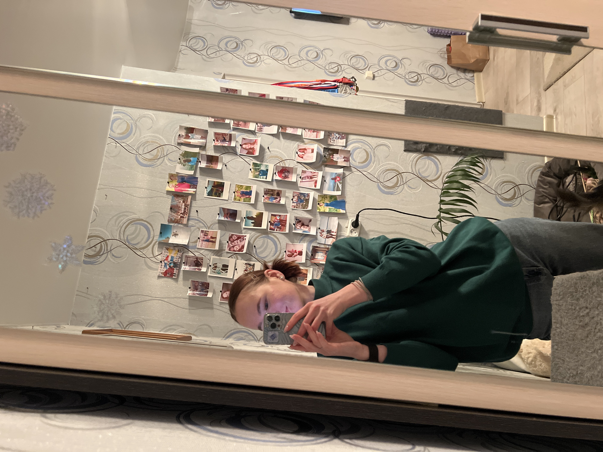 A woman is reflected in a mirror. She is holding a smartphone and appears to be taking a picture. The background wall is decorated with a swirling pattern wallpaper and a collection of small photographs arranged in a grid. A houseplant is partially visible on the right. The image was taken in Chaikovsky, Russia.