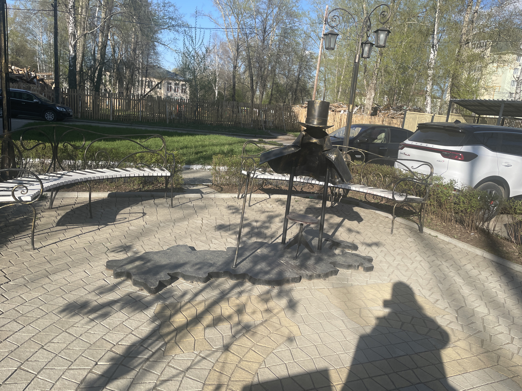 The image shows a public space in Chaikovsky, Russia, featuring a bronze sculpture of Anton Chekhov sitting on a bench. The sculpture is surrounded by three semicircular benches. A lamppost with three lamps is positioned behind the statue. The ground is paved with interlocking concrete blocks. In the background, there is a wooden fence, green grass, trees, and two cars. The photograph includes the shadow of the person taking the picture.