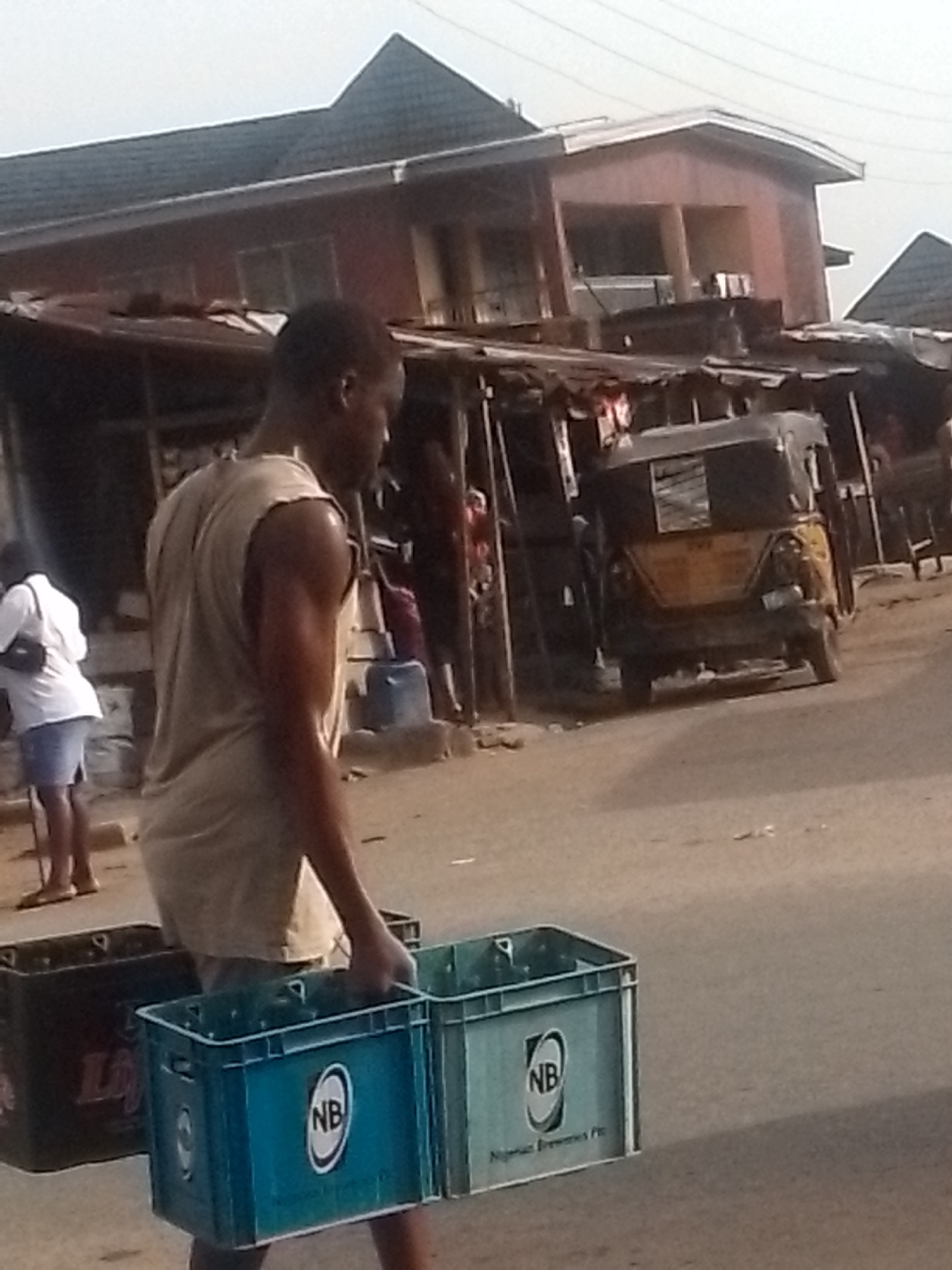 A man carries three crates of beverages in the street. Another person, a woman, stands in the background. A three-wheeled motor vehicle is visible in the background. The location appears to be a commercial area. The city and country are unknown.