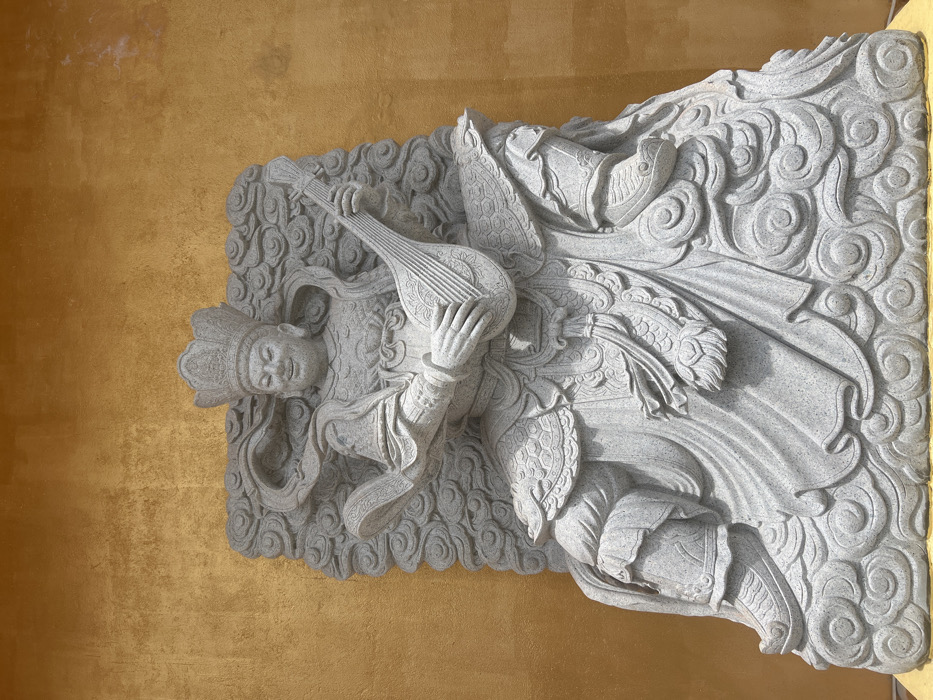 A grey granite statue of a regal figure is centered against a textured golden-yellow wall in Đăk Mar, Vietnam. The figure, depicted seated with a benevolent smile and closed eyes, wears an elaborate crown and intricate robes with armor-like elements. It is actively engaged in playing a stringed, lute-like instrument with both hands. The figure is carved upon and surrounded by stylized cloud patterns that form its base and backdrop within the overall stone carving. The stone exhibits a speckled, light grey texture.