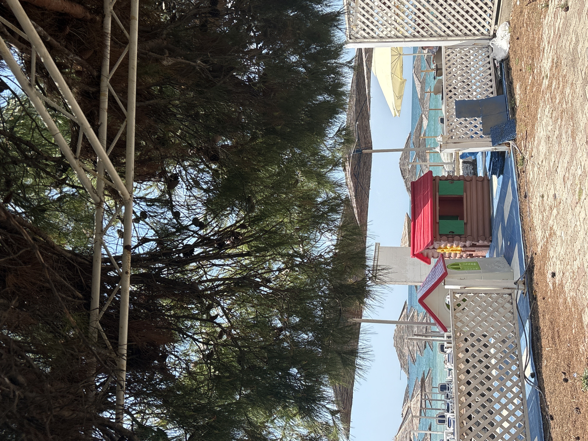 The image depicts a beach scene in Bar, Montenegro, viewed from underneath a canopy of pine trees. In the foreground, a white lattice fence separates the viewer from a play area with two small, colorful playhouses. One playhouse resembles a log cabin with green shutters and a red roof, while the other is a white structure with a red roof and a green window. The ground in the play area is blue and tiled. Beyond the fence and play area, rows of beach umbrellas and lounge chairs are set up on the sand, leading to the turquoise waters of the Adriatic Sea. The sky is clear and blue. The scene appears to be a resort or a public beach area.