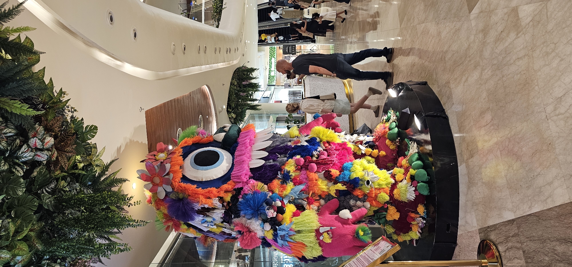 The image, taken in Denpasar, Indonesia, shows an indoor scene in a shopping center or mall. The main focus is a large, colorful artistic sculpture or display that resembles a whimsical creature or monster with a large eye. This object is placed in a shiny, black rounded container.
Several people are visible in the background. A man wearing a dark shirt and jeans is leaning over the display, likely taking a photo of the sculpture. Nearby, a woman in a light-colored outfit is walking with her back towards the camera. Further in the background, near the railing of an upper level, there are several other individuals, appearing as small figures.
The environment includes retail stores, escalators, and lush greenery, including what appears to be a vertical garden. The interior has a modern design with a curved ceiling and sleek surfaces. The floor is made of light-colored tiles.