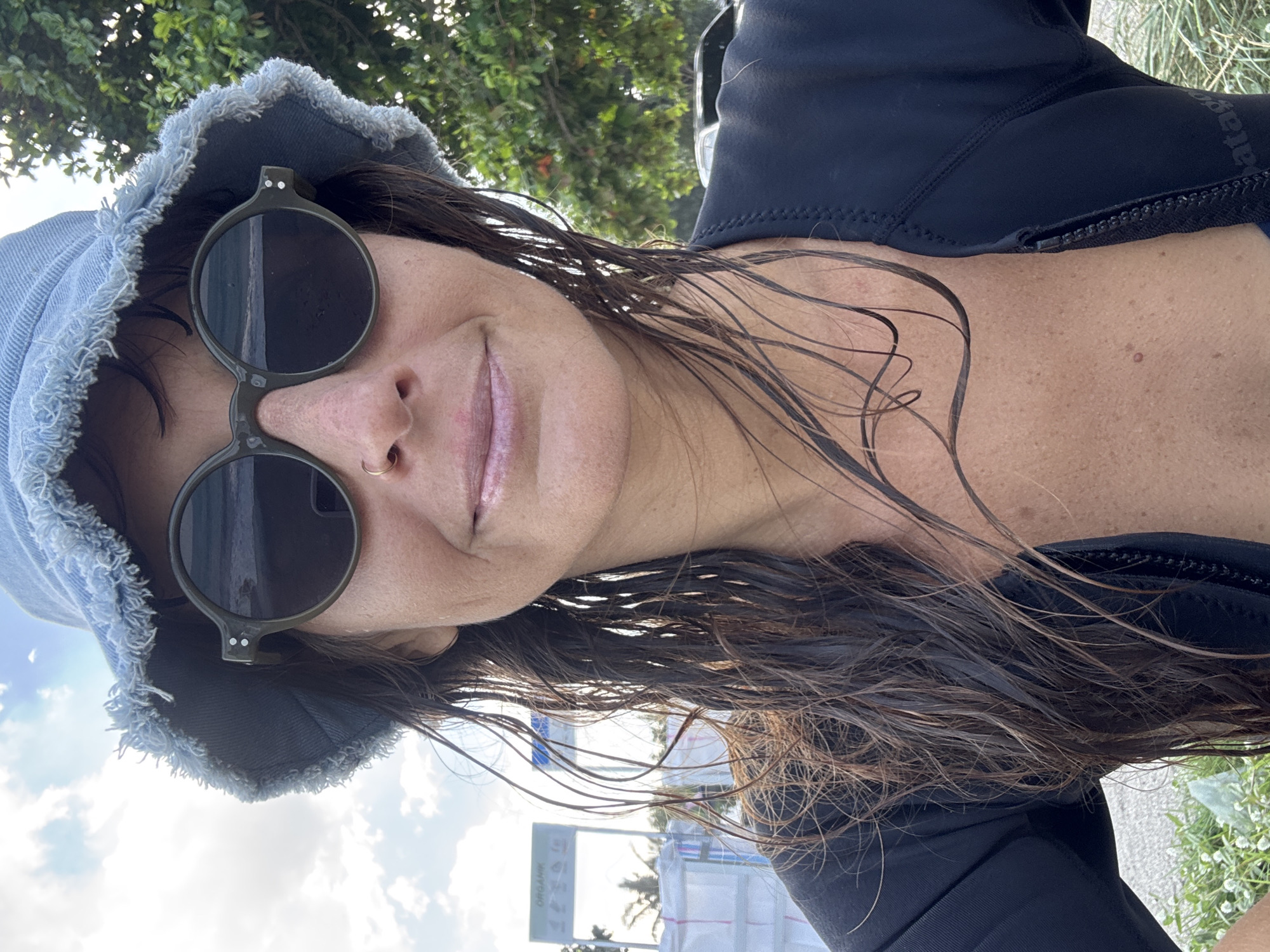 In an outdoor, full-body, eye-level, medium shot, a woman with dark, wet hair, wearing a black wetsuit and a light blue denim bucket hat, poses for a selfie. She is wearing round, dark sunglasses, and has a gold nose ring. The background shows a cloudy sky, trees, and some white structures with a sign that reads "ORGANIK." The scene appears to be a beach or coastal area.