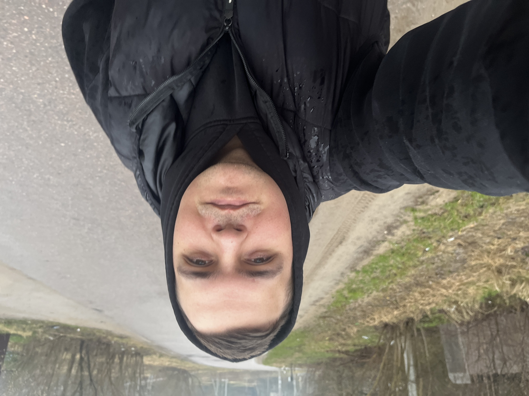 The image shows a person in a black hooded jacket standing on a paved road. Water droplets are visible on the jacket, indicating it may be raining or recently rained. The background features roadside vegetation, with dry grass and some green patches. Trees are visible in the distance, suggesting a possibly suburban or semi-rural location. The setting appears to be Раздолье, Russia.