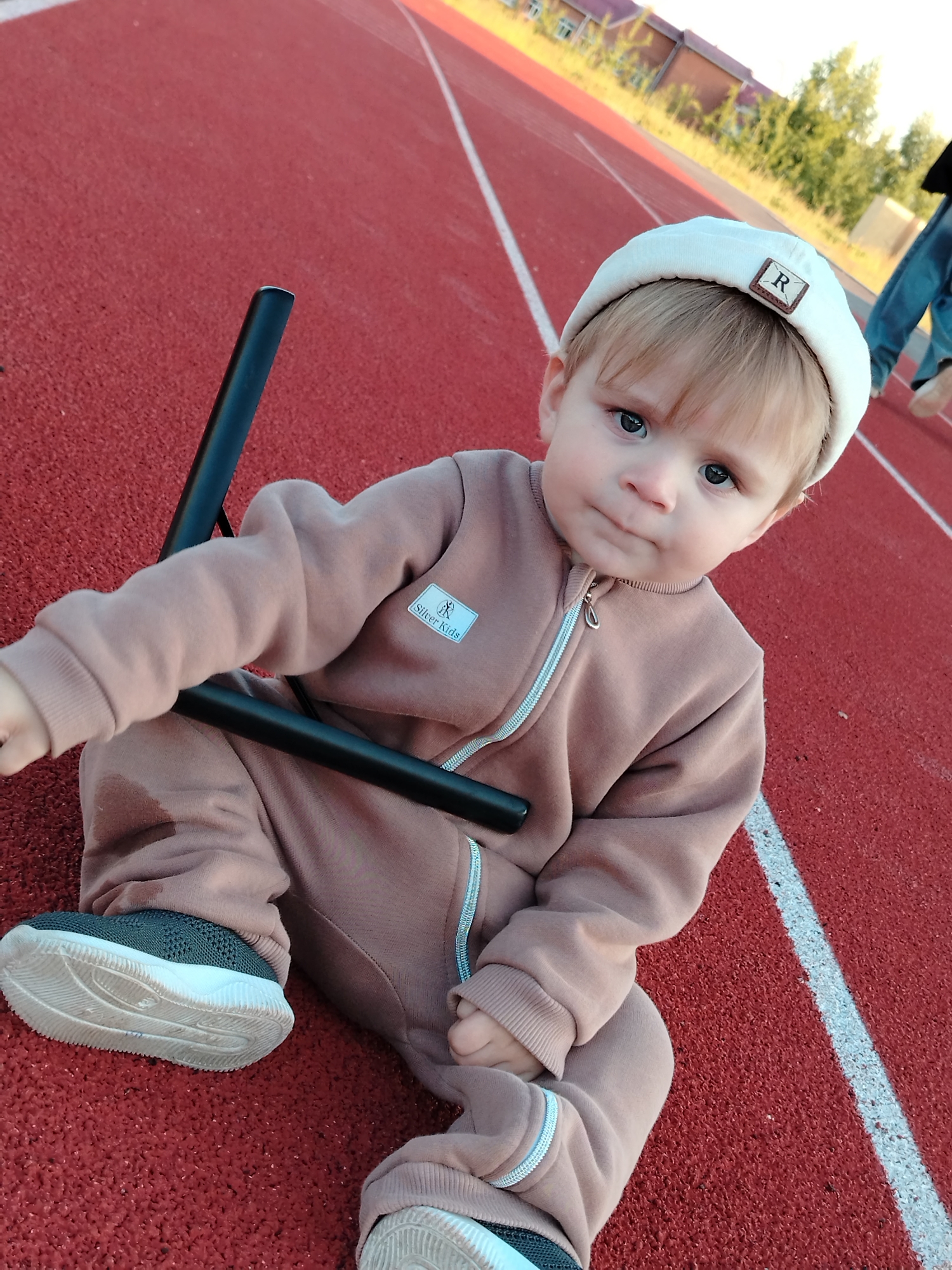 A young child, appearing to be a toddler or baby, is captured in a close-up, slightly low-angle shot, sitting on a red running track. The child, likely male, has light blonde hair and dark eyes, and is wearing a light cream-colored beanie with a brown square patch featuring the letter 'R'. He is dressed in a light brown or taupe full-body suit with a silver zipper down the front and a label on the left chest that partially reads "Silver Kids". On his feet are grey and white sneakers. The child is holding onto a black, cylindrical object, possibly a part of a stroller or a piece of equipment. His expression is neutral, looking directly at the camera with a slight pout.
The setting is an outdoor sports track, identifiable by the distinctive red, textured surface and white lane markings. In the background, out of focus, are green trees, some dry grasses, and a red-brick building, suggesting the location is likely a school or community sports ground in Noviy, Russia. The lighting indicates late afternoon or early evening, with warm sunlight illuminating the scene. An adult's legs, clad in blue jeans, are partially visible on the right side of the frame, walking away.