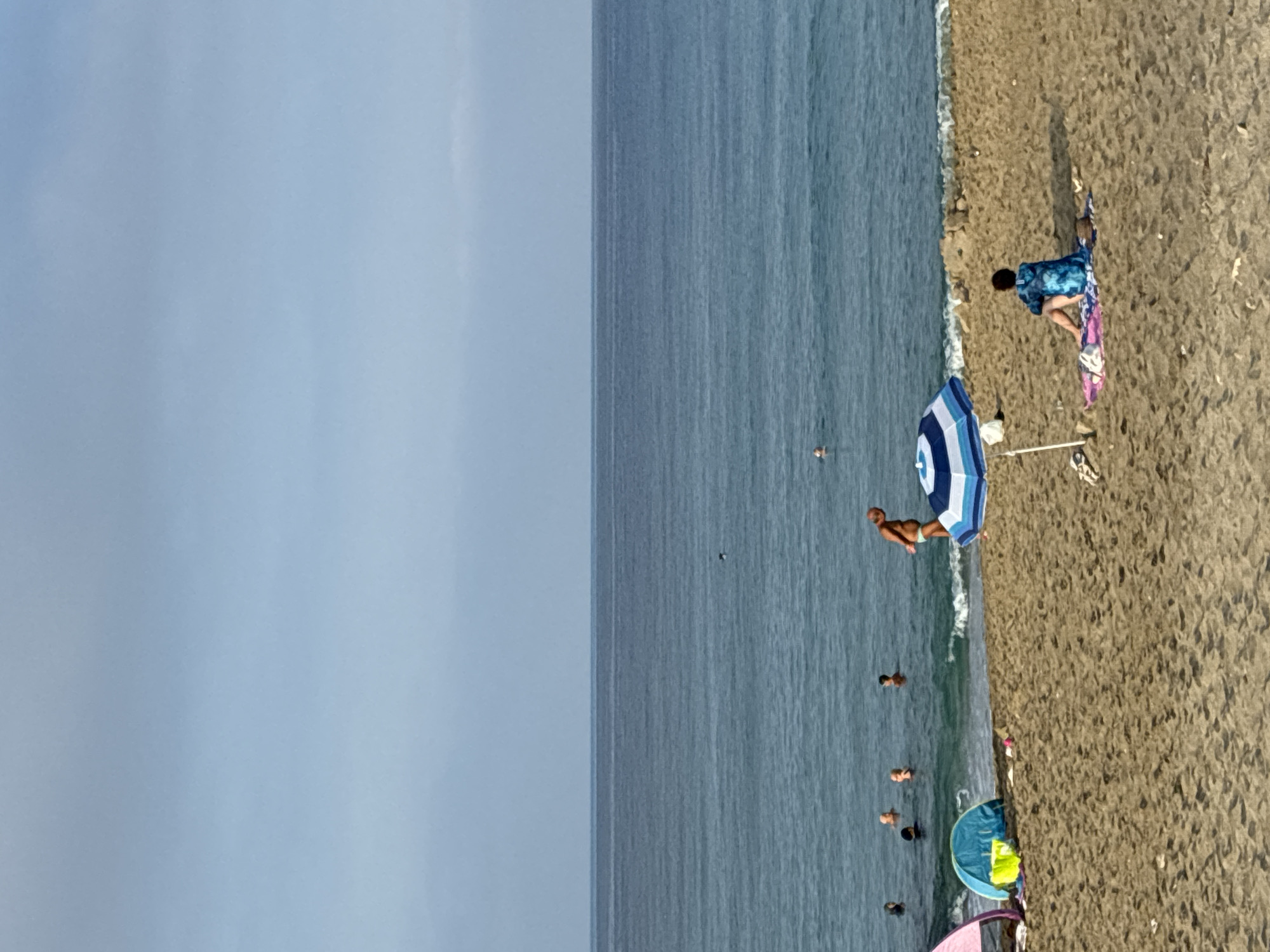 The image depicts a beach scene with people in the water and on the sand, under a clear sky with a haze near the horizon. The sea is calm with gentle waves reaching the shore. Several individuals are visible in the water, some appearing to swim or wade. On the beach, an adult male in light-colored swimming briefs stands near the water's edge. Further on the sand, a person is seated facing away from the camera, wearing a blue tie-dye garment and resting on a blanket. A blue and white striped umbrella is set up next to them. In the foreground, the sandy beach shows footprints and some scattered items, including a towel with a colorful pattern. To the left, a pink beach tent and a blue beach tent are set up, with a yellow bucket nearby. The overall impression is of a leisurely day at the beach. The location is confirmed to be Alanya, Turkey.