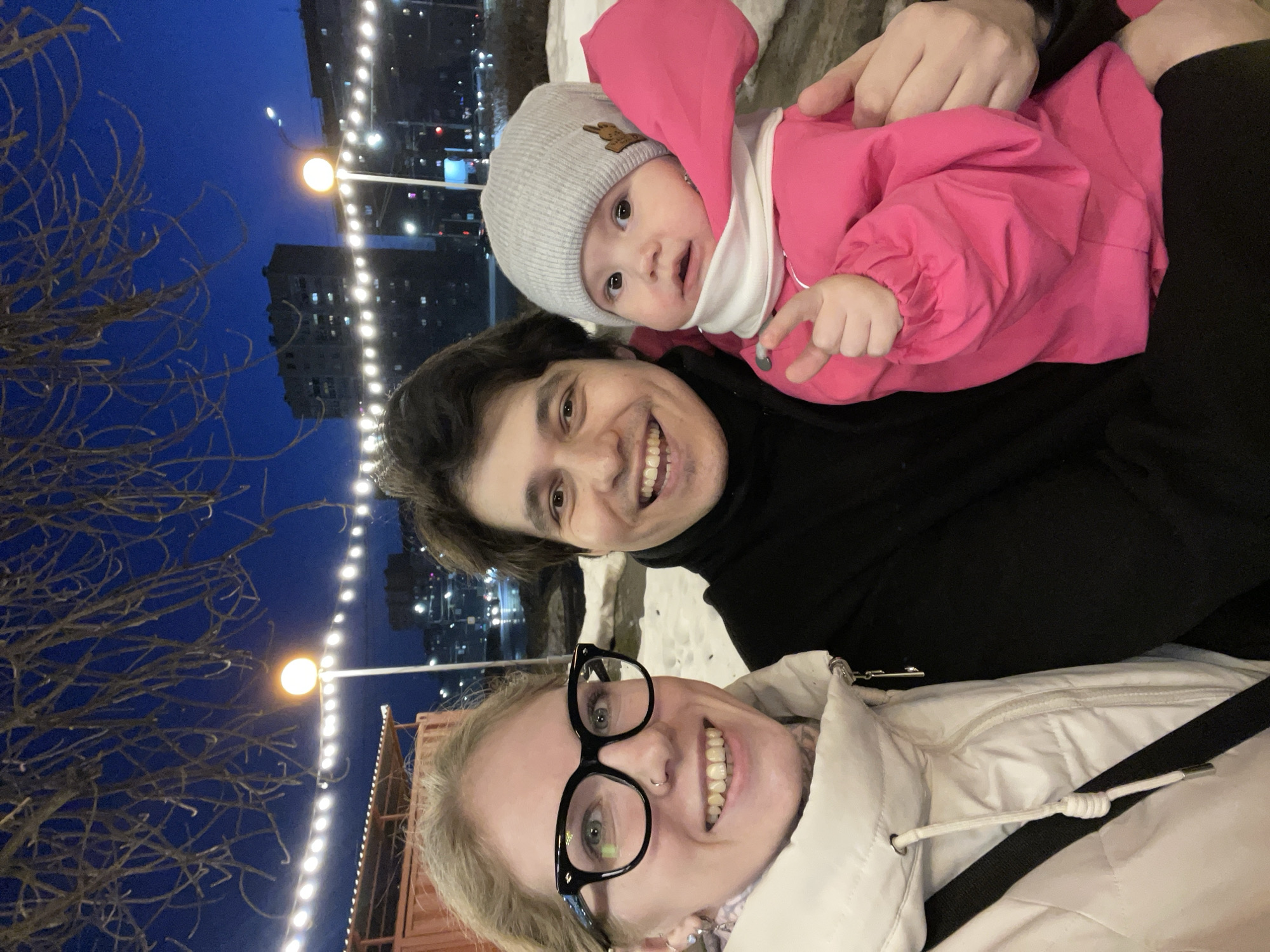 A close-up, rotated, outdoor selfie depicts three individuals: a woman, a man, and a baby. The woman, positioned at the bottom of the frame, wears glasses and a white jacket. Above her is a man in a black top. A baby wearing a grey hat and pink jacket is being held by the man. The baby is pointing with its right hand.
The background features a dark blue evening sky, streetlights, bare trees, and a tall building with illuminated windows. This scene is in Izhevsk, Russia.