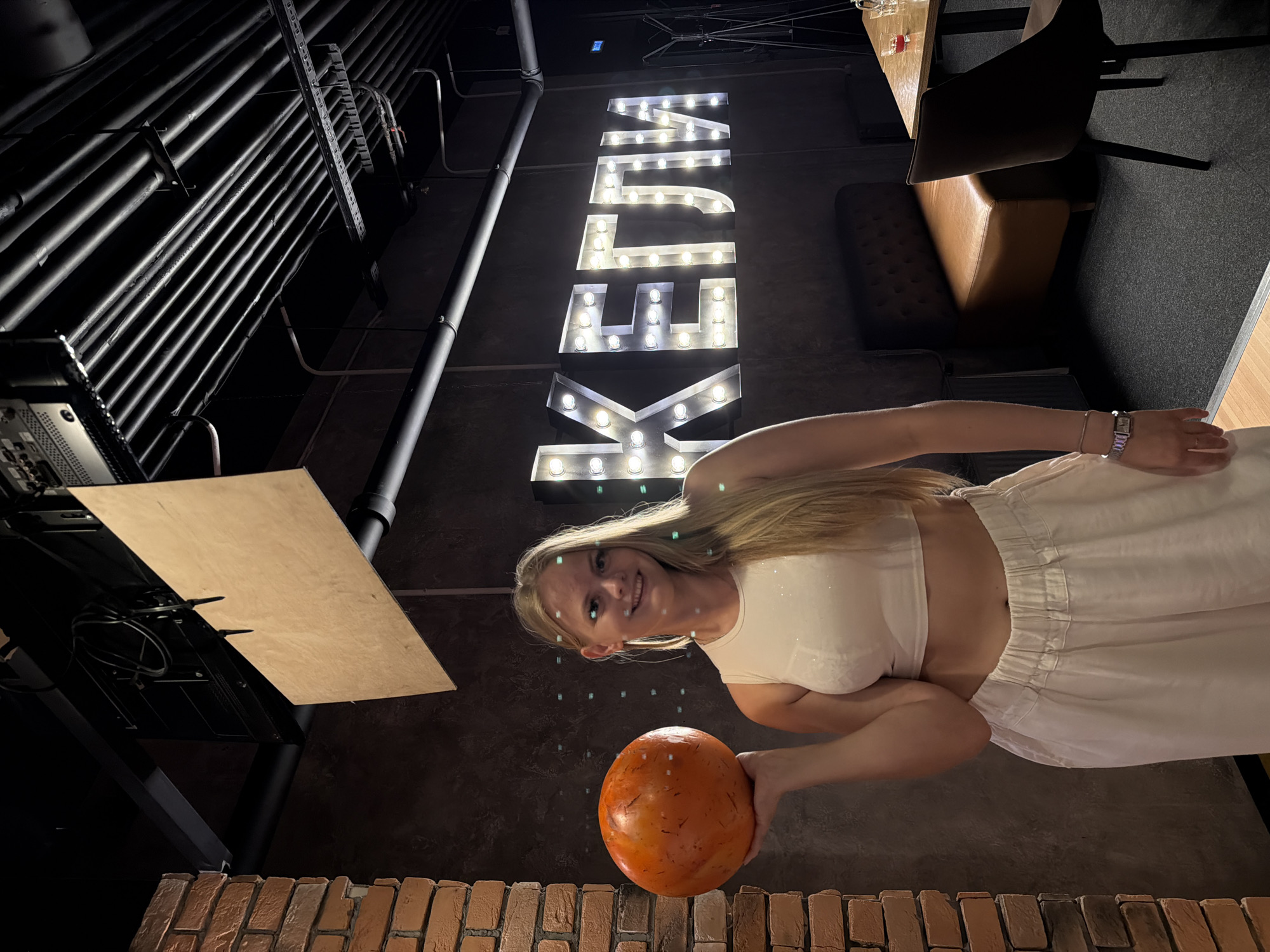 This image shows a young woman standing in an indoor setting with industrial-style decor. She is smiling and holding an orange, round object, possibly a bowling ball. The woman is wearing a light-colored crop top and a flowing white skirt, revealing her midriff. She has blonde hair and is looking towards the camera.
The background features a large, illuminated sign with block letters that spell out "KELLA" in a retro, marquee style with many light bulbs. To the left of the sign, there are numerous dark pipes running along the wall. Further to the left, a wooden panel is propped up, and some electronic equipment is visible beneath it. In the background on the right, there is a glimpse of a leather booth seat and a table with some glasses. The floor appears to be a dark textured material. The lighting suggests it is nighttime or an indoor setting with artificial lighting.
The overall impression is a casual, possibly entertainment-focused environment, given the bowling ball and the prominent sign. The woman appears happy and relaxed. The setting has a gritty, urban aesthetic with exposed pipes and brickwork visible at the bottom of the frame. The location is contextually identified as Izhevsk, Russia.