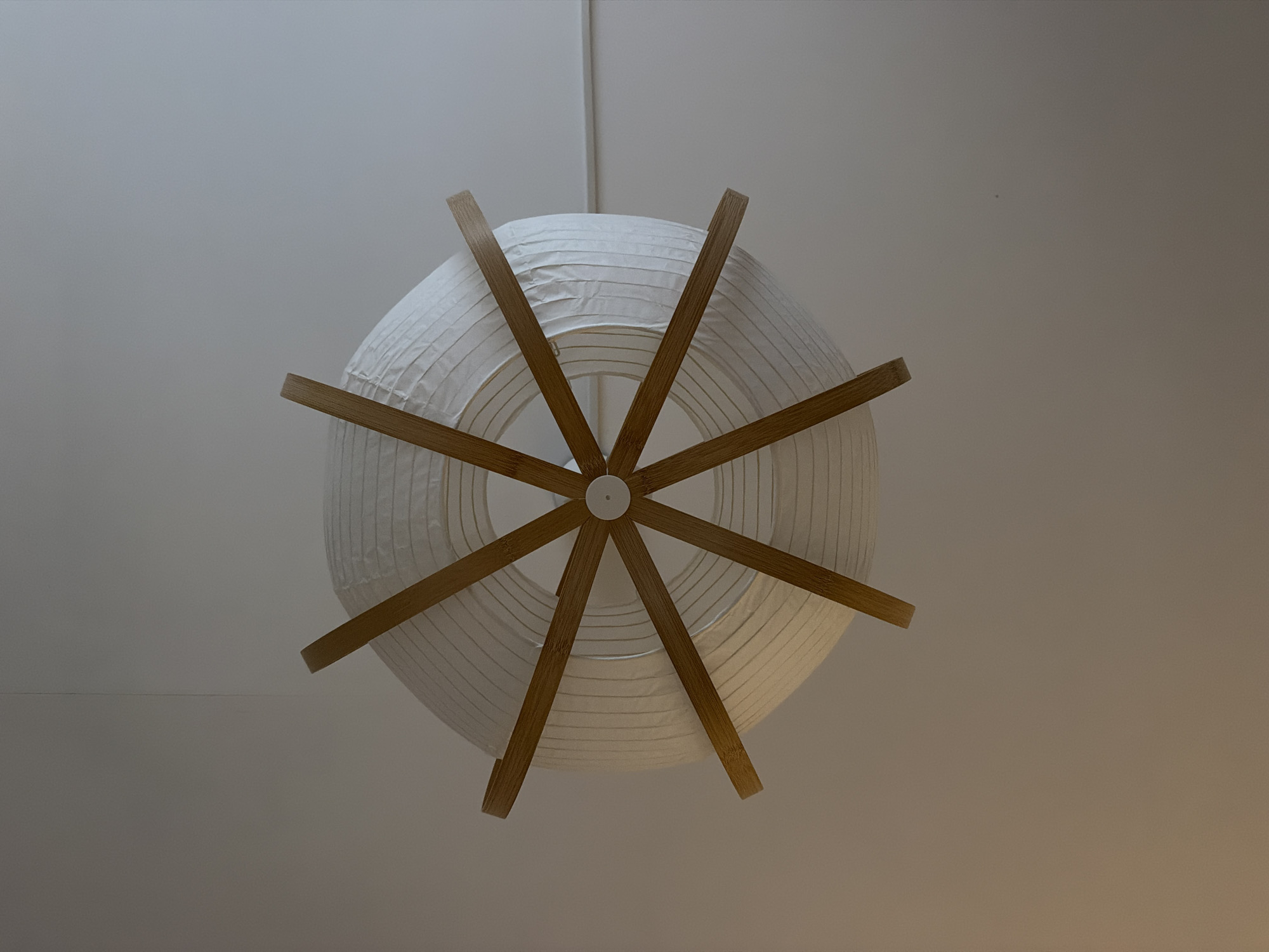 This is a top-down, medium shot of a white paper lantern light fixture hanging from a white ceiling. The lantern is cylindrical with a thin, grooved texture and is supported by thin wooden slats that extend from a central point in a starburst pattern. The wooden slats are a light brown color and appear to be made of bamboo. The ceiling is smooth and off-white, with a subtle shadow cast by the lantern. The lighting is soft and diffused, suggesting an indoor environment. No people or other objects are visible in the frame.