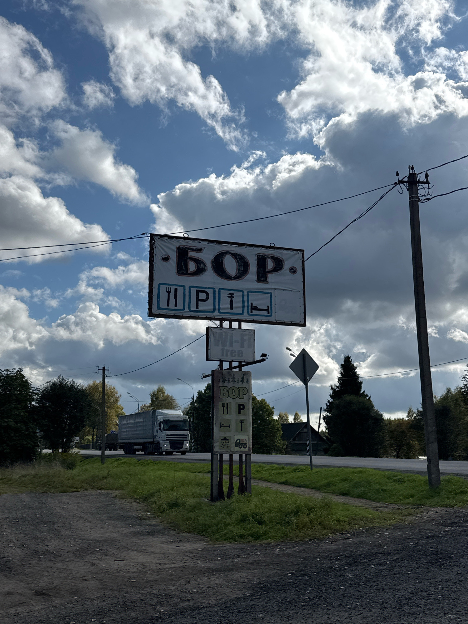 In Novyi Berezai, Russia, a roadside scene unfolds under a dramatic sky filled with scattered cumulus clouds against a blue backdrop. A large, weathered sign, featuring Cyrillic text that appears to read "БОР" and symbols for a fork and knife, a bed, and possibly a toilet, stands prominently in the foreground. Below this main sign, a smaller sign displays "Wi-Fi free" and another "БОР" sign with similar icons. A white semi-trailer truck drives on a paved road to the left of the frame. Dense green trees line the right side of the road, interspersed with a dark green evergreen tree. A wooden structure, possibly a house, is visible behind the trees. A power pole with multiple wires stretches across the upper right portion of the image. The ground in the foreground consists of a gravelly, dusty surface.