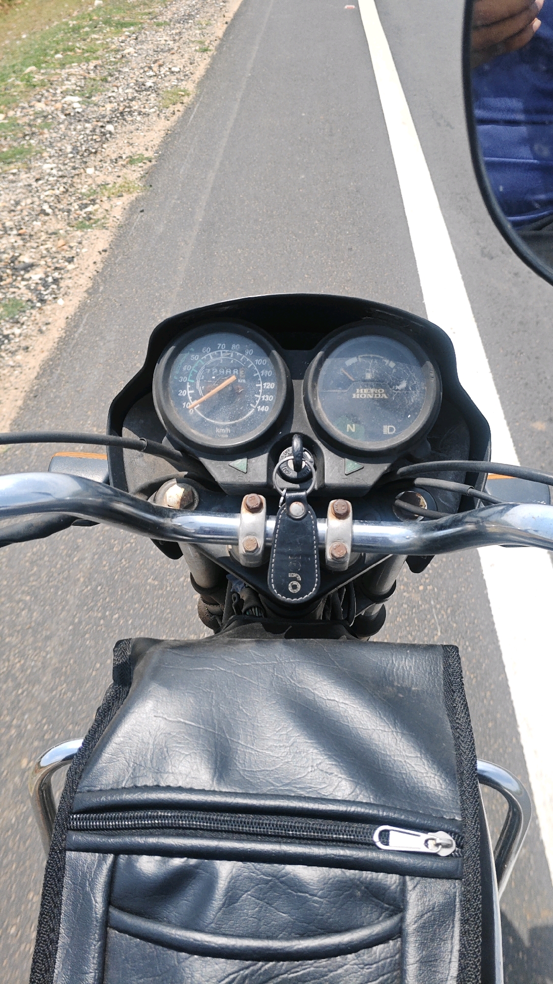 The image shows a first-person perspective from a motorcycle traveling on a paved road. In the foreground, the motorcycle's handlebars and a black leather bag with a zipper are visible. Behind these, the motorcycle's dashboard features two circular meters, one likely a speedometer showing approximately 20 km/h, and the other an indicator for gear position. A key with a black leather fob is attached to the dashboard. The road has a white line marking its center and a gravelly shoulder on the left. The environment suggests an outdoor setting in a rural or semi-urban area, consistent with Kotta Jadadevi, India, during daytime. A reflection in the rearview mirror shows a person wearing a blue garment.