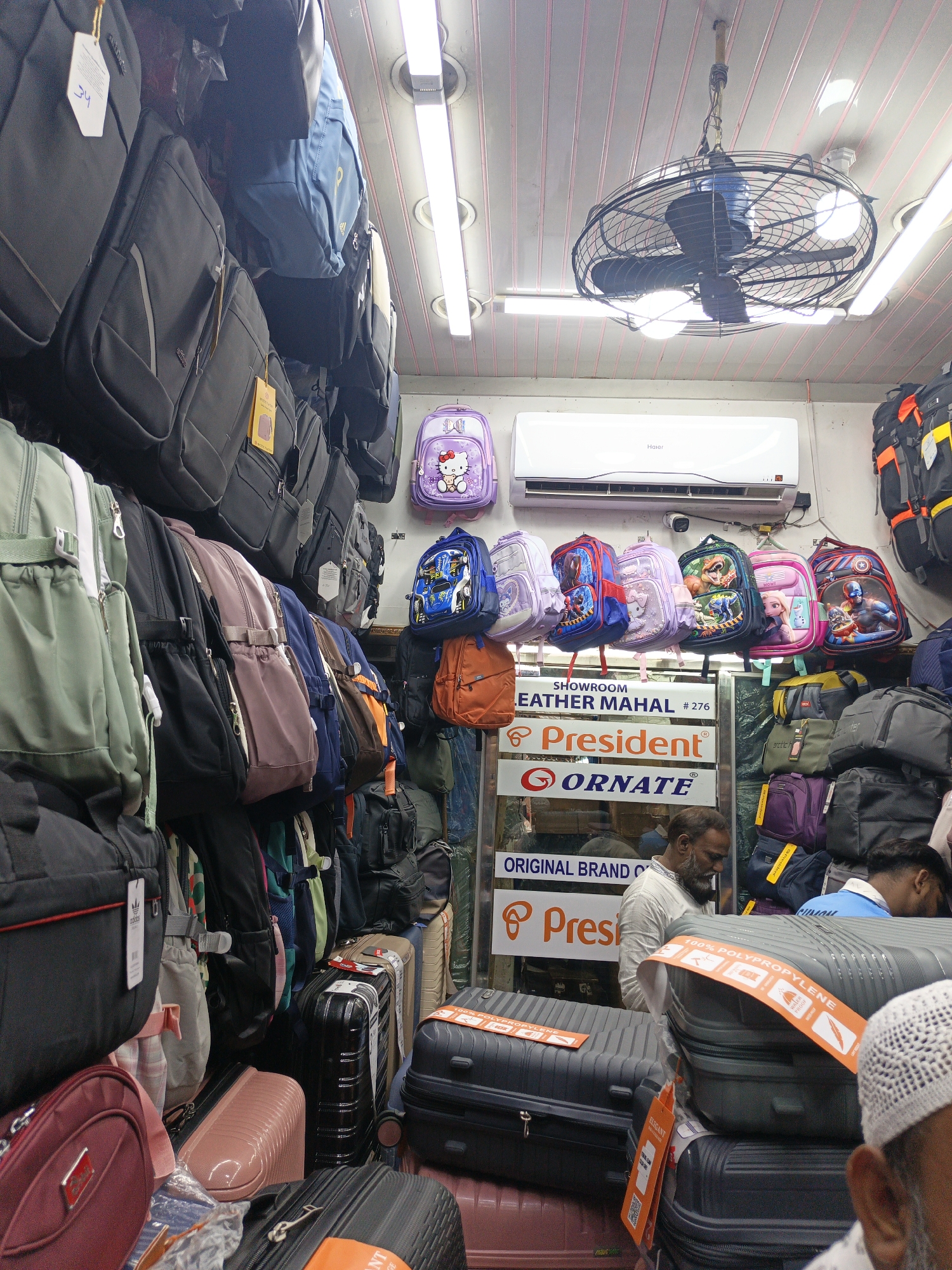 This indoor market scene, located in Dhaka, Bangladesh, depicts a densely packed bag and luggage shop. The setting is characterized by an overwhelming display of merchandise, with various types of bags covering walls, shelves, and floor space. Overhead fluorescent lights and a black caged ceiling fan with integrated lights illuminate the shop. A white Haier air conditioner unit is mounted high on one wall, indicating a commercial environment that likely manages temperature.
Numerous backpacks of diverse colors and designs hang from hooks and racks. These include children's backpacks featuring popular characters such as Hello Kitty, Spider-Man, dinosaurs, Elsa from Frozen, and Captain America, alongside adult backpacks in solid colors like black, grey, green, blue, pink, brown, and maroon. On the shop floor, hard-shell suitcases, primarily grey and black, are stacked. Some suitcases are secured with orange straps that read "100% POLYPROPYLENE."
At least two men are present. One man with a beard and a light-colored shirt stands behind a display of suitcases, his gaze directed downwards. Another person's head, wearing a white prayer cap (kufi), is partially visible in the bottom right corner, obscured by luggage. The atmosphere appears to be that of a typical, busy retail operation.
Visible text includes store signage for "SHOWROOM LEATHER MAHAL #276," along with brand names such as "President®" and "ORNATE®," and the phrase "ORIGINAL BRAND C." Product tags display text like "ELEGANT" and "arctich," and a price tag shows "34." The air conditioner is branded "Haier."
