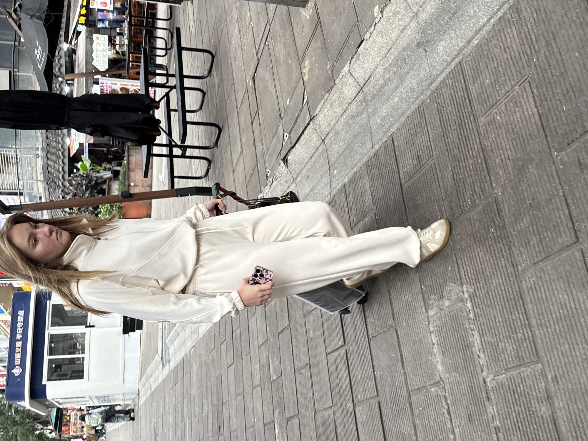 A woman is walking along a sidewalk in Yuzhong District, China. She is wearing a white tracksuit and sneakers, holding a smartphone. An umbrella is nearby. The sidewalk is made of grey pavers. There are shops and cafes in the background.