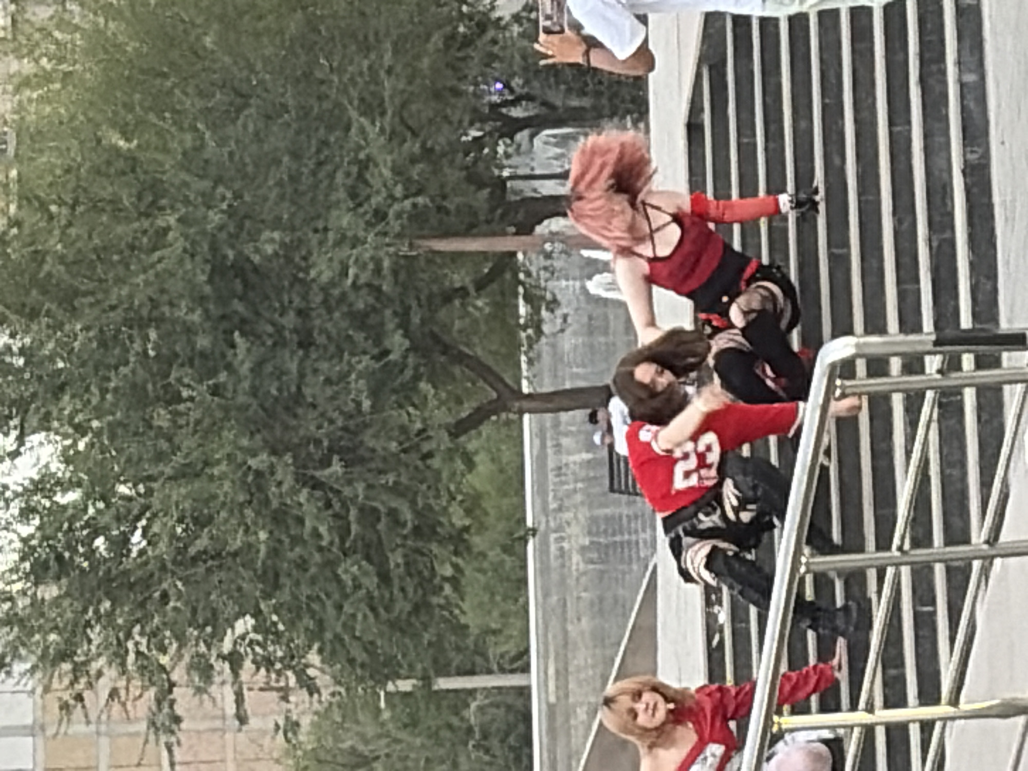 In an outdoor urban setting, three young women are captured in motion on a tiered concrete staircase surrounded by trees. One woman on the left, with blonde hair, wears a red off-the-shoulder top and red pants, her body angled towards the viewer. In the center, a woman with dark brown hair is dressed in a red jersey with the number "23" and patterned pants, leaning forward with her hands near the stairs. To the right, a woman with bright pink hair wears a red tank top with black straps and black shorts, her body twisted as if dancing or performing an action. Another person, only partially visible on the far right, is wearing a white t-shirt and holding a phone, appearing to record the scene. The staircase is a prominent feature, with metal railings in the foreground. The background includes trees and a building.