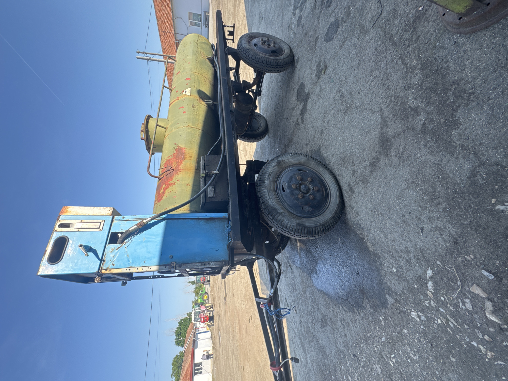 The image depicts an old, rusty fuel truck or tanker parked on an asphalt surface. The sky is clear blue. The truck has a green tank and a blue cab. The overall condition of the vehicle is deteriorated. The location is Sarnevo, Bulgaria. Other vehicles, including tractors, are visible in the background. A building with a red roof is also present.
