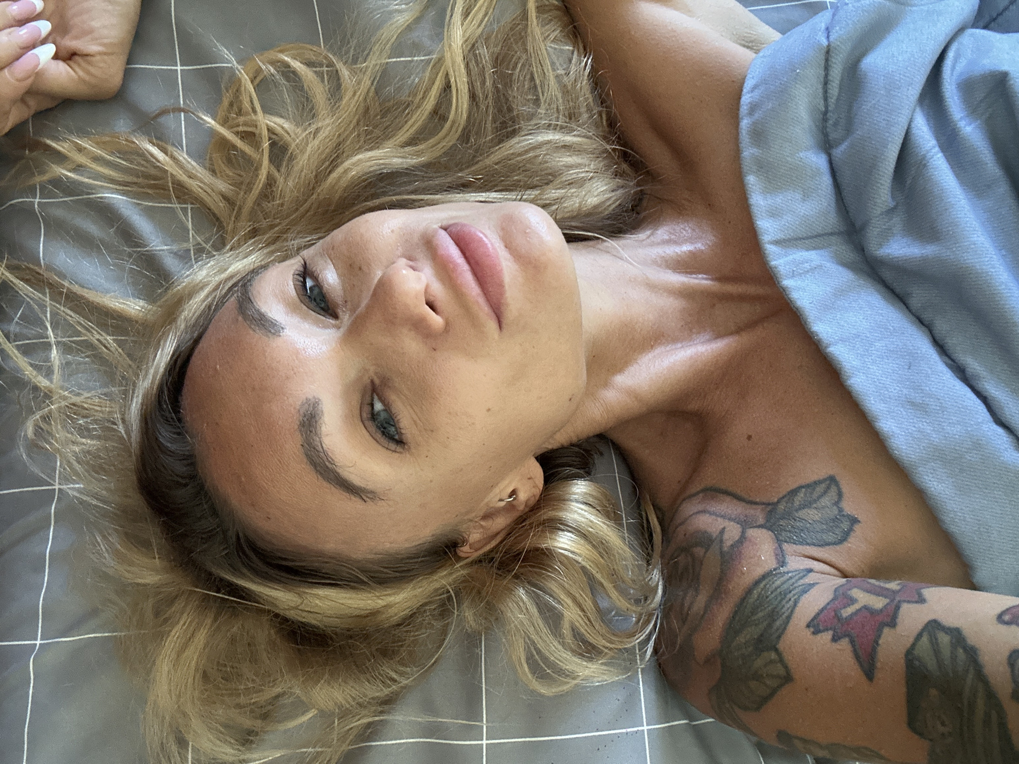 The image is a close-up, overhead shot of a woman lying on a gray, checkered bedspread. She has blonde hair, blue eyes, and is wearing light makeup. Her head is slightly tilted, and she is looking towards the camera with a relaxed expression. A light blue blanket is draped over her lower body and shoulder. There is a tattoo of flowers on her left arm. A hand with French manicured nails is visible in the upper right corner of the frame. The scene appears to be indoors, possibly a bedroom. There is no discernible information that would place this image in Mũi Né, Vietnam.
