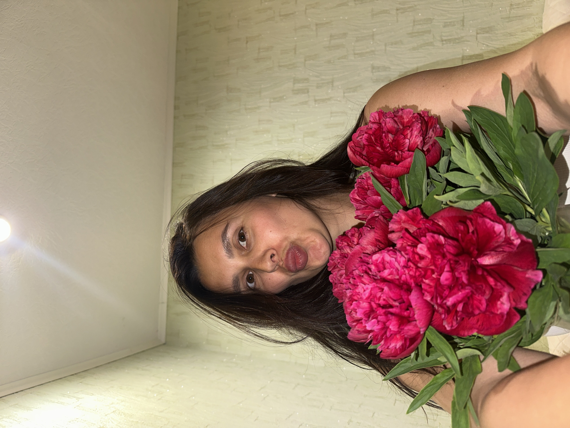 A woman with dark hair is holding a bouquet of red peonies. She is looking at the camera with a pursed-lips expression. The background consists of a wall with a textured pattern and a ceiling with a light source. There is no verifiable information about the location being in the city of Chaikovsky, Russia.