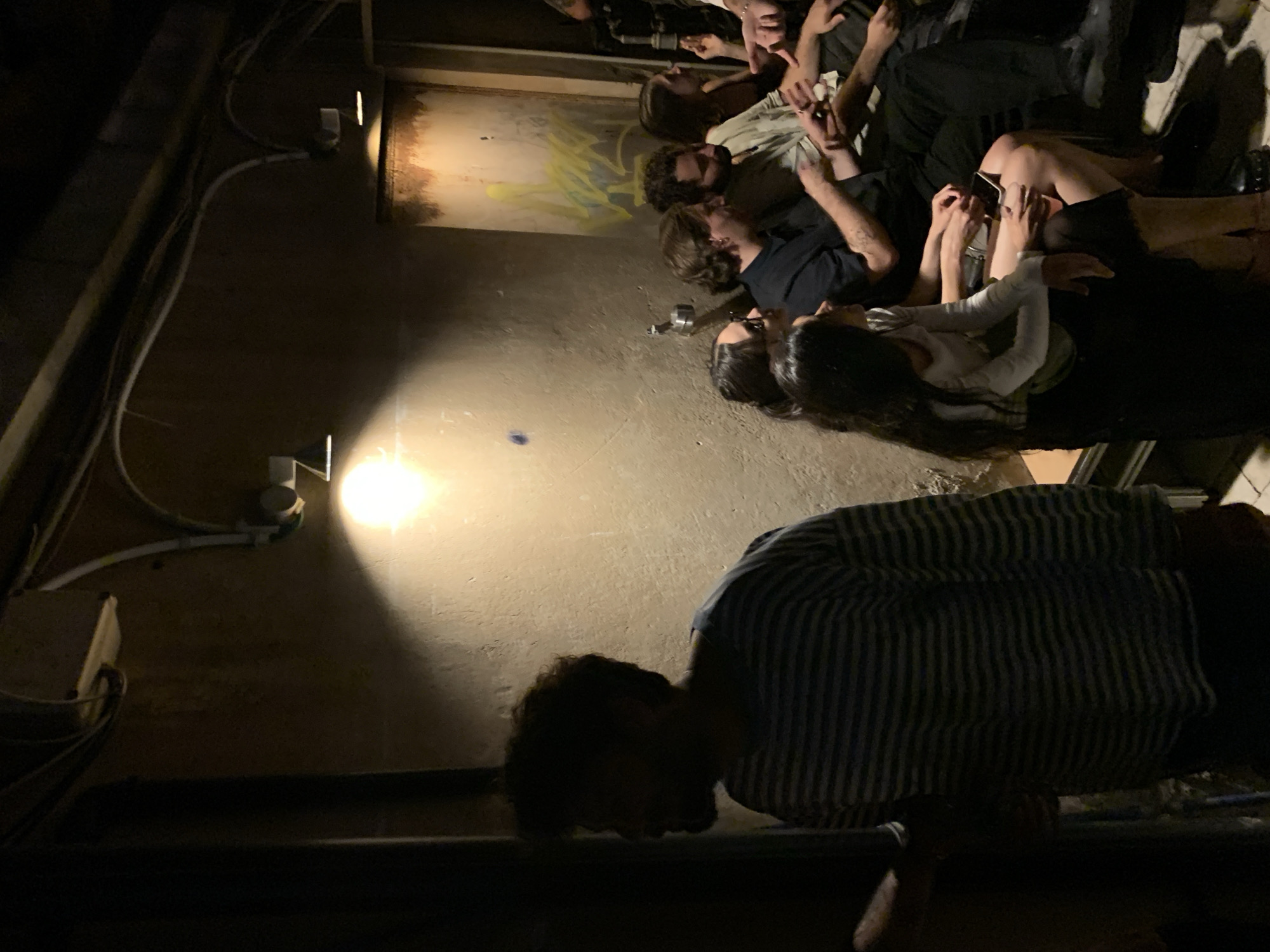 The image depicts an indoor scene with a group of people seated on a bench against a textured wall. The lighting is dim and dramatic, with a bright overhead light casting long shadows. The wall appears to be concrete or stucco, with some signs of wear and tear, and a graffiti tag is visible on the right side.
In the foreground, a person wearing a horizontally striped long-sleeved shirt is partially visible, silhouetted against the wall. To the right, a group of individuals are seated, engaged in conversation and interaction. One person, a woman with dark hair and glasses, is looking towards the left. Next to her, a man with light brown hair is wearing a dark shirt. Further to the right, a man with dark curly hair and a beard is wearing a light-colored shirt. Beside him, a woman with long dark hair is looking forward, and her right arm is raised. Another woman with dark hair is seated next to her, wearing a white long-sleeved shirt and a black skirt, holding a drink in her left hand. The people are seated on a bench that appears to be made of wood or a similar material.
The environment suggests a casual, possibly dimly lit venue like a bar, club, or lounge. The overall impression is of an intimate gathering. Given the context of Tel-Aviv, Israel, this scene could be taking place in any number of urban nightlife establishments.