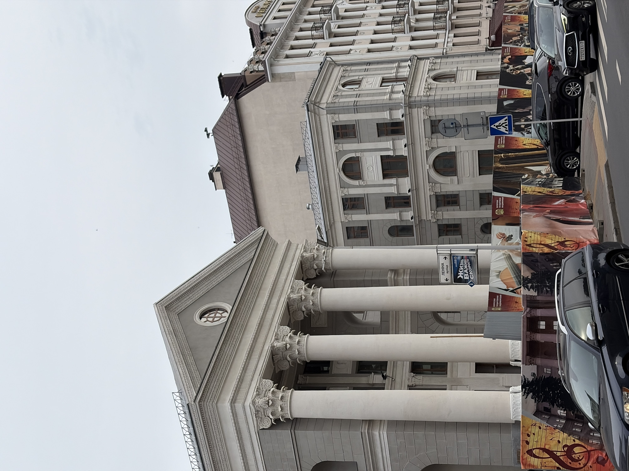 This detailed image captures a street scene in Minsk, Belarus, focusing on a collection of buildings under an overcast sky.
In the foreground, a multi-paneled construction hoarding, adorned with musical notes, architectural sketches, and images of string instruments, spans the width of the frame, partially obscuring the lower sections of buildings and parked cars.
Dominating the left side is a grand, classical-style building with a grey facade and white ornamental details. It features a prominent portico supported by four tall, fluted white Corinthian columns. Above the columns, a pediment houses a circular oculus window with a cross-shaped mullion. The building's upper facade also displays decorative cornices and moldings. Behind this building, the roofline of an older, plainer beige-colored structure with a reddish-brown metal roof and visible chimneys is apparent.
To the right of the classical building, another multi-story building with a light beige facade and white decorative elements stands. Its lower visible floors feature arched windows, while the upper floors have rectangular windows with small black wrought-iron balconies. A distinctive sign is partially visible on its upper right side, reading "HOTEL ЕВРОПА" (Hotel Europa).
The street in front of the hoarding is asphalt, with white pedestrian crossing markings visible on the right. Several vehicles are present:
* On the left, parked parallel to the curb, is a dark blue Volvo V70 or XC70 station wagon, facing towards the right. It has silver roof rails and window trim.
* Behind the Volvo, partially hidden by the hoarding, is a black SUV, possibly a Hyundai Santa Fe.
* On the right, closer to the viewer and positioned partially on the pedestrian crossing, is a black Infiniti QX70 SUV, facing left. Its license plate, "7272XI-7", is clearly visible.
A vertical pole rises from the sidewalk, displaying multiple signs. The uppermost is a blue directional sign with white arrows pointing left and right, indicating "ЛЕНІНА вуліца" (Lenina Street). Below it is another blue sign with white text, "ЖИЗНЬ ВАЖНЕЕ СКОРОСТИ" (Life is more important than speed), with a downward-pointing arrow. Further down, a blue square sign with a white pedestrian crossing symbol is affixed. No individuals are visibly present in the scene. The overall lighting is soft and diffused, characteristic of an overcast day.