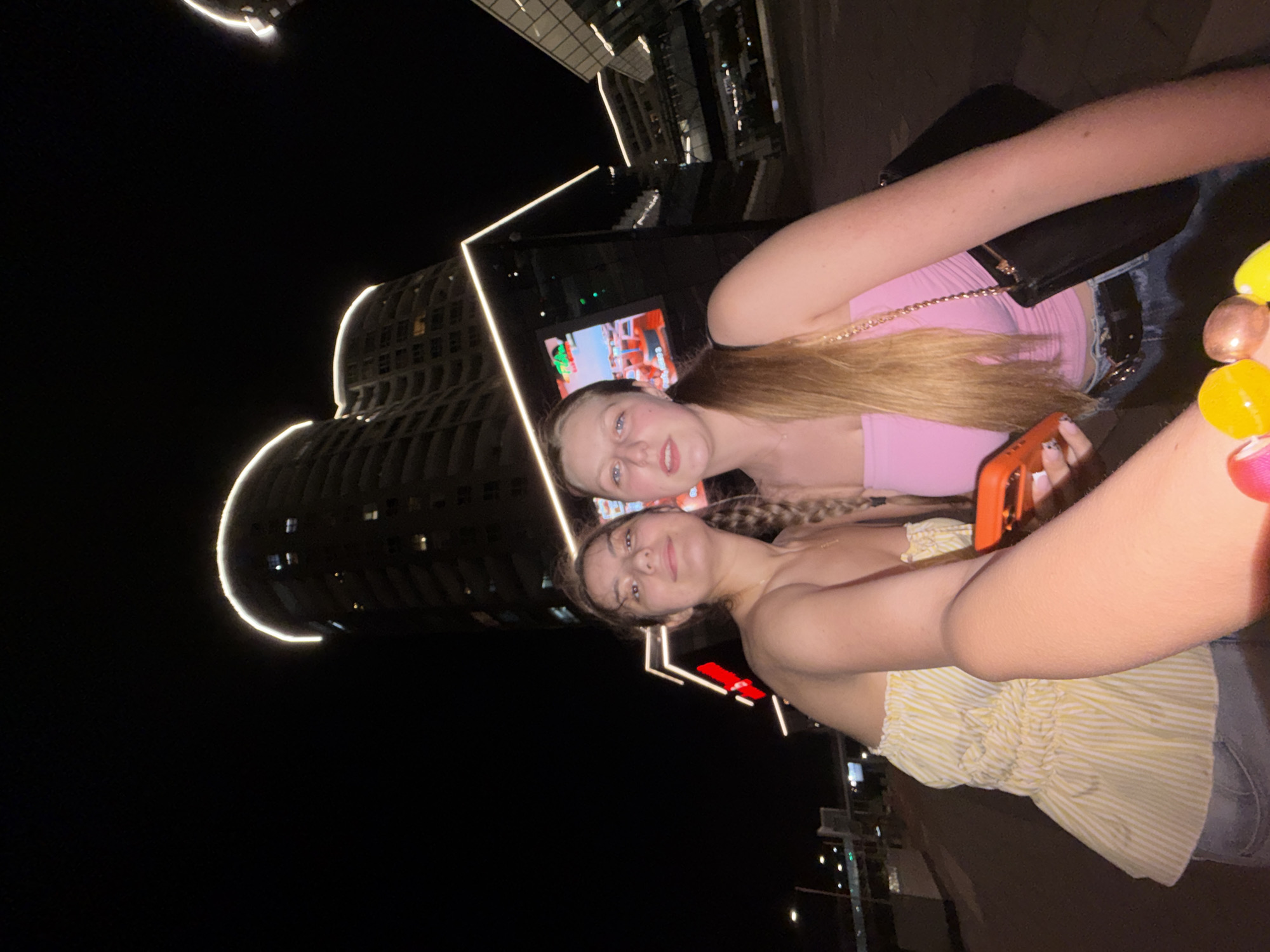 This nighttime photograph captures two young women in an urban setting in Burgas, Bulgaria. The image is taken at an angle, likely a rotated selfie or group photo.
In the foreground, a young woman with brown hair, styled in braids, wears a yellow and white striped strapless top. A colorful beaded bracelet adorns her right arm. Next to her, another young woman with long blonde hair wears a pink strapless top. She holds an orange-cased smartphone in her hand, suggesting she might be taking the photo. Both women look directly at the camera with neutral to slightly smiling expressions.
The background is a dark night sky, illuminated by modern urban architecture. Distinctive buildings with sleek designs are visible, outlined by bright white LED light strips that trace their contours. One building in particular has a prominent curved shape, dramatically lit. Behind the women, a large digital screen or billboard displays vibrant, colorful imagery. The overall scene suggests an evening outing in a contemporary city environment. No discernible text is clearly visible on the screen or buildings.