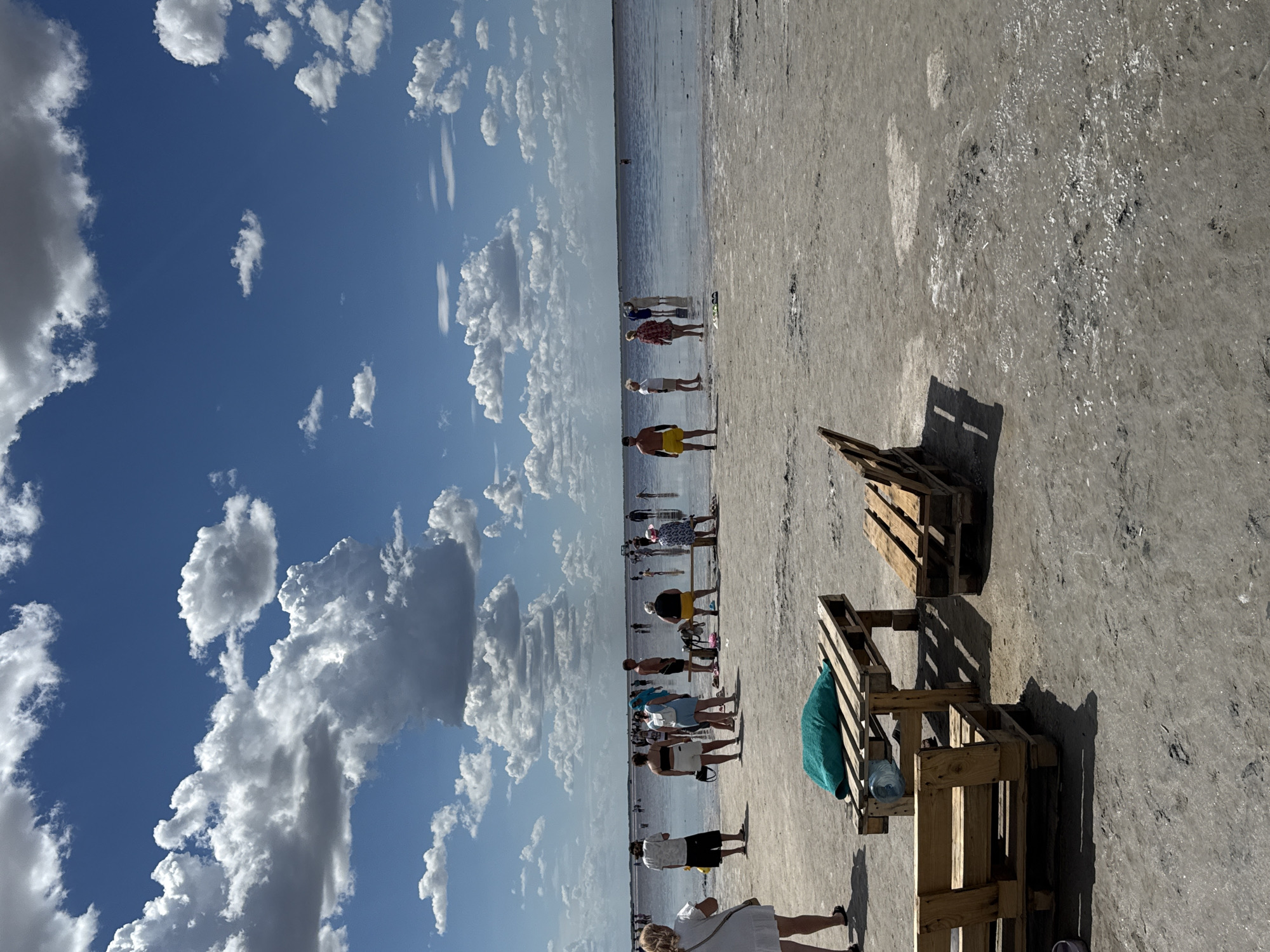 The image depicts a sunny day at a salt lake beach in Бурсоль, Russia. The sky is a vibrant blue, dotted with fluffy white clouds. In the foreground, there are several makeshift wooden furniture pieces constructed from pallets, serving as benches and a table. A teal towel and a water bottle are visible on the table. The ground is a light-colored, dry salt crust with scattered debris.
In the mid-ground and background, numerous people are enjoying the water and the beach. They are dressed in casual summer attire, with some wading in the shallow, reflective water, while others stand on the shore. The lake stretches out to the horizon, appearing calm and shallow. The overall scene conveys a sense of leisure and recreation at a unique natural destination.