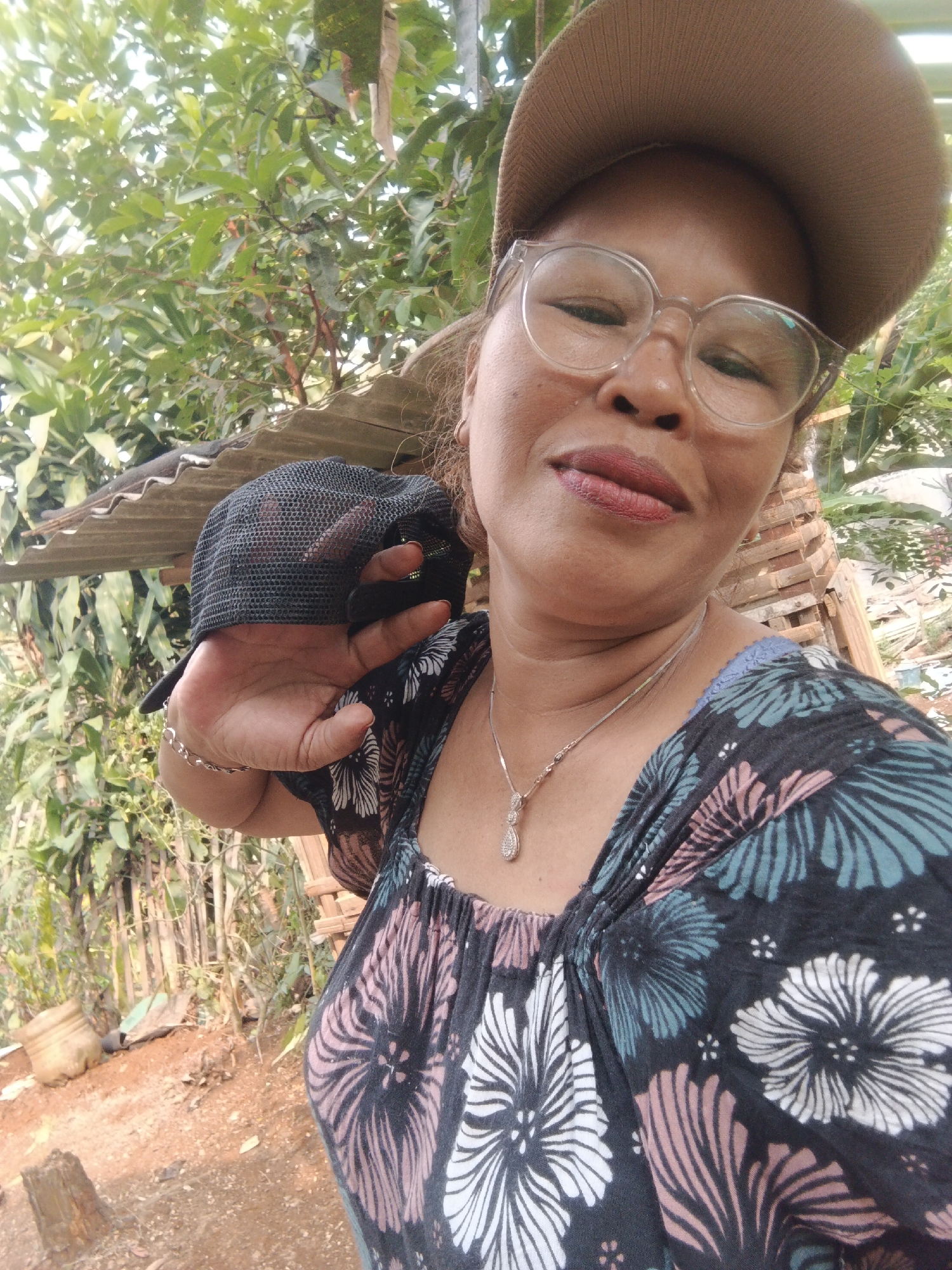 A woman, wearing a brown baseball cap and clear-rimmed glasses, is taking a selfie outdoors. She has on a black t-shirt with a floral pattern of pink, white, and teal flowers. A silver necklace with a teardrop pendant is visible on her neck. Her left hand, adorned with a silver bracelet, is raised, obscuring part of her face and the camera. The background consists of lush green foliage, a corrugated metal roof structure, and wooden elements, suggesting a rural or semi-rural setting. The ground is reddish-brown dirt with some scattered debris, including a plastic bottle. The overall lighting suggests daytime. The image appears to have been taken in Warungkiara, Indonesia.