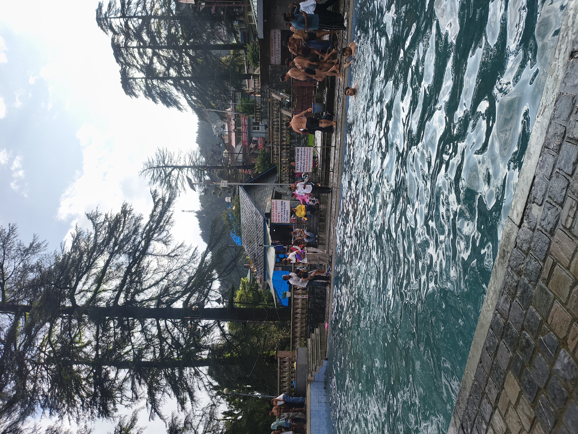 The image depicts a public swimming pool in Bhagsu, India, on a bright, overcast day. The pool is filled with clear, turquoise water, with ripples indicating movement. Several individuals are visible in and around the pool. In the foreground, two young males are swimming, with their heads and upper bodies visible above the water. Along the edge of the pool, a group of men, some shirtless and wearing shorts, are standing and walking. One man is reaching into a blue bucket. Further back, several people, including men, women, and children, are seated on benches or standing near a building with a blue awning. One person in a yellow jacket is seated on the far left. Behind the pool area, there are tall pine trees and buildings, suggesting a hillside location. The sky is visible, with clouds and patches of bright light. The ground around the pool is paved with dark stones. Signs with text in Hindi are posted on the wall behind the pool.
