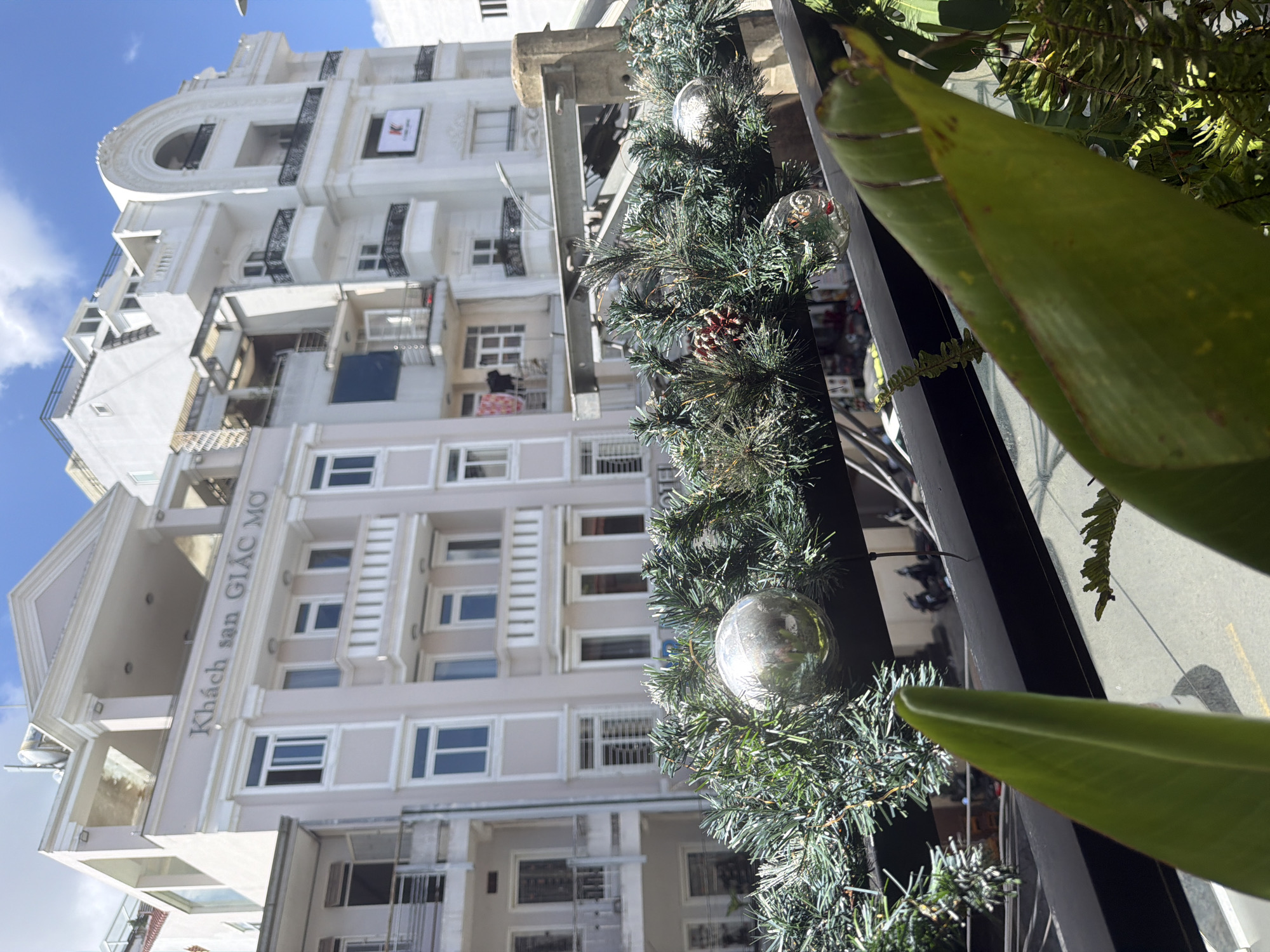 The image depicts a street scene in Da Lat, Vietnam, focusing on the facade of a building with signage indicating "Khách sạn GIÁC MỌ." The building is multi-story and painted in light colors, featuring numerous windows and balconies. In the foreground, a decorative garland of pine branches and Christmas ornaments is visible, suggesting a seasonal display. The street below shows parked motorcycles and a pedestrian, indicating normal urban activity. The architecture is a blend of modern and possibly older European styles, characteristic of many buildings in Da Lat. The sky is blue with scattered clouds.
