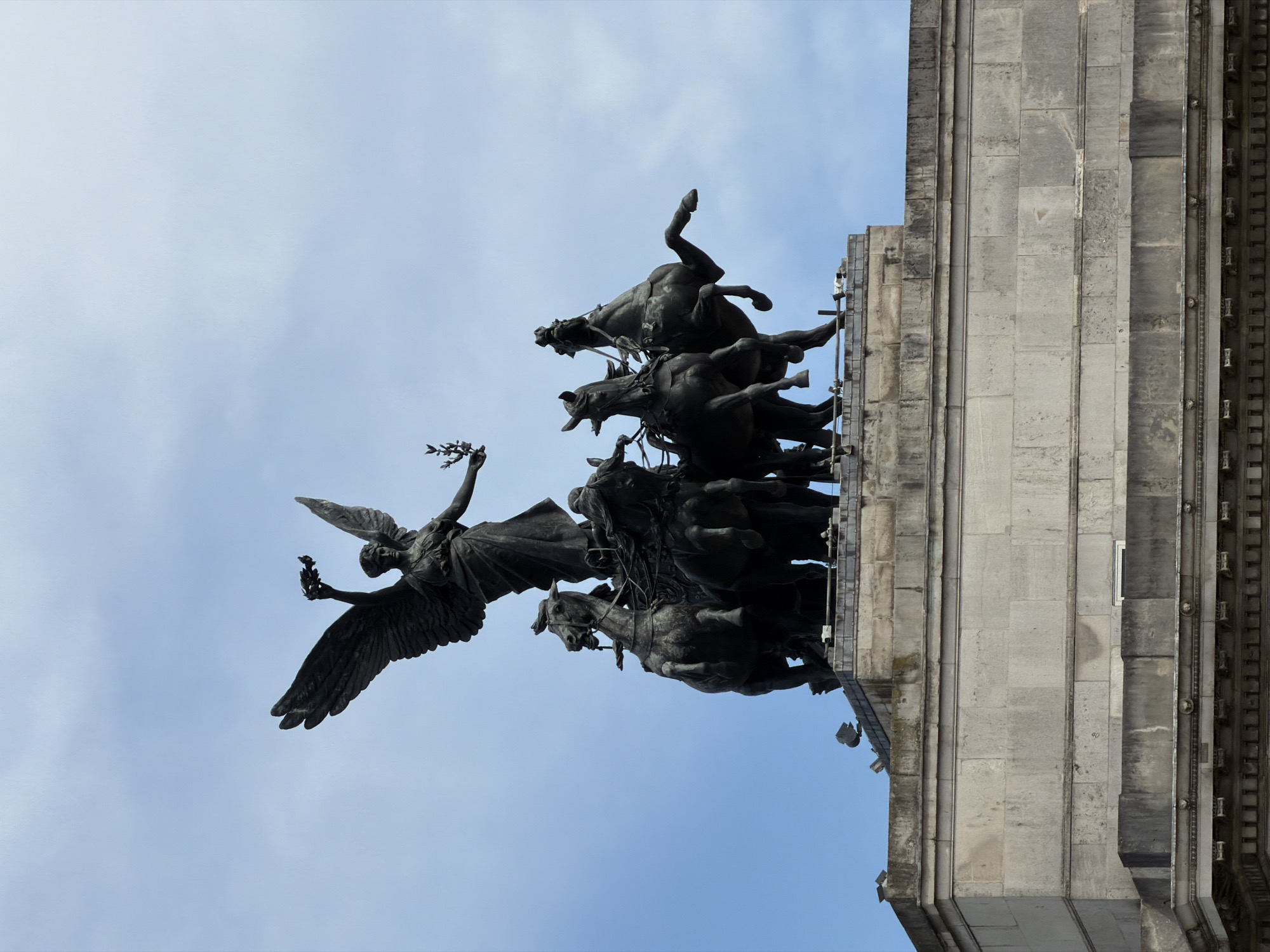 This image captures a magnificent bronze sculpture group atop a grand stone building, viewed from a low angle against a bright, light blue sky with subtle cloud formations, indicating daytime.
The central subject is a dynamic quadriga, a chariot pulled by four powerful horses depicted in various states of rearing and motion, conveying immense energy and forward thrust. Several male figures, likely charioteers or soldiers, are also part of the group, though their details are somewhat obscured by the dark bronze material.
Leading this dramatic procession is a majestic winged female figure, possibly representing Victory or Peace. She holds a laurel wreath or branch in her outstretched left hand, symbolizing triumph or honor, while her wings are broadly spread, enhancing the sense of flight and momentum. The dark, aged bronze of the sculpture contrasts sharply with the pale sky.
The sculpture is mounted on a substantial stone edifice, likely a triumphal arch, which occupies the right side of the frame. This structure is built from large, light-colored stone blocks and features classical architectural elements, including prominent pilasters, cornices, and detailed decorative carvings that suggest historical significance. The stone shows signs of weathering and age.
The overall impression is one of grandeur, power, and triumph. Given the location context of London, United Kingdom, this artwork is identifiable as the Quadriga on top of the Wellington Arch at Hyde Park Corner. No visible text is discernible on the sculpture or the building.
