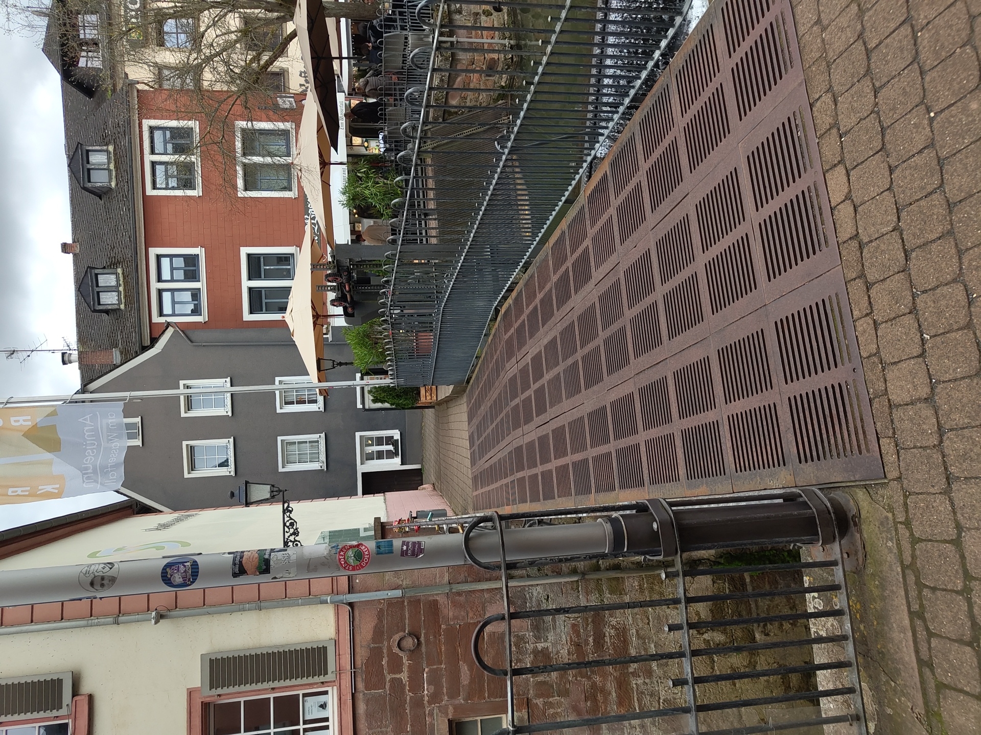 The image captures a view of Saarburg, Germany, showing a street scene alongside a waterway. In the background, there are multi-story buildings painted in various colors such as grey and red, with visible windows and roofs. Overlooking the waterway are people under umbrellas, likely seated at a restaurant. A metal railing runs along the water's edge. In the foreground, a metal grate covers part of the walkway, alongside brick pavers. A pole covered in stickers and another metal fence are also visible in the foreground.