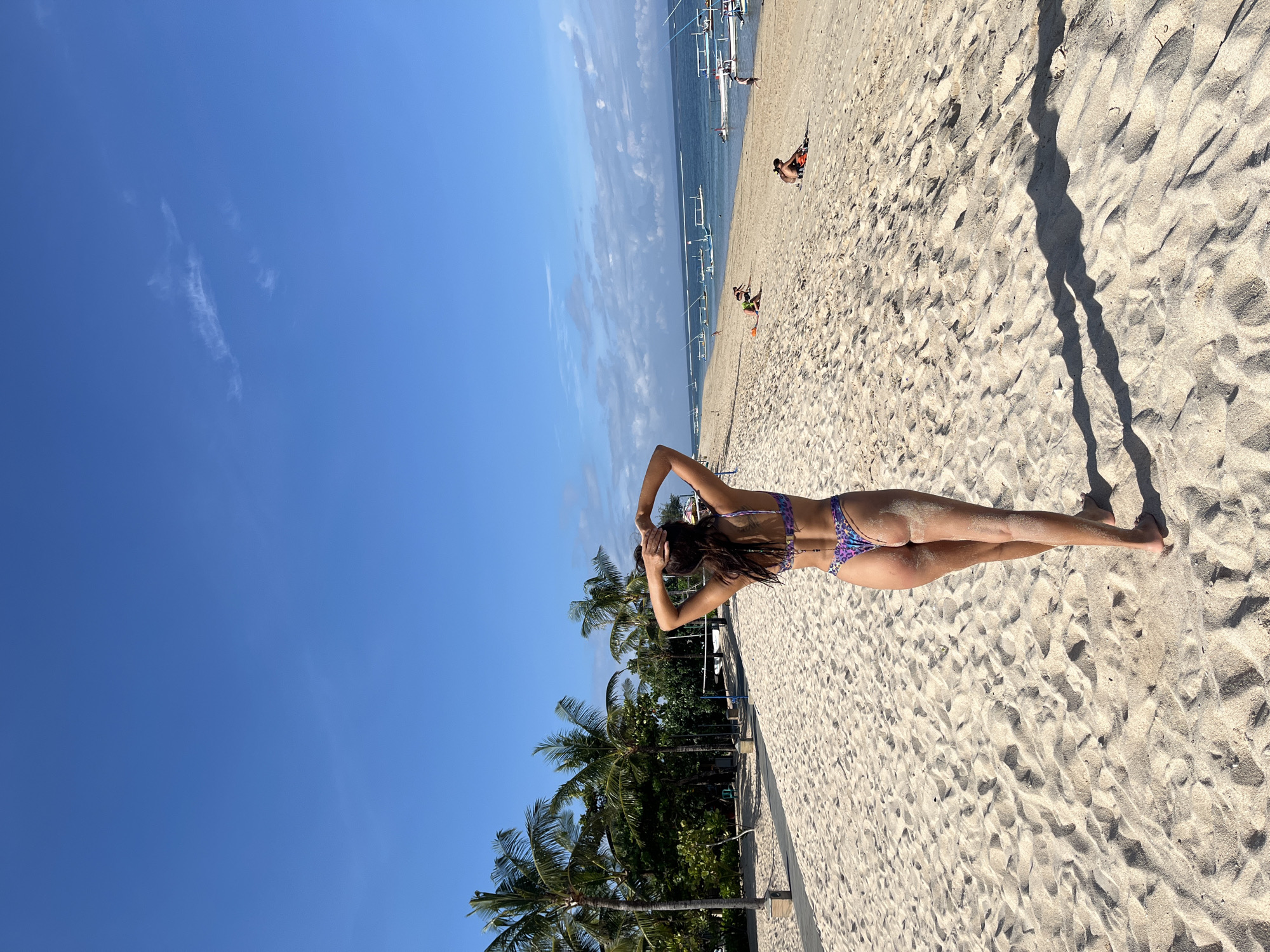 A woman wearing a multicolored bikini is lying prone on a sandy beach under a sunny sky. Her arms are raised, hands behind her head. Palm trees are visible near the lower left of the frame, along with some fencing. In the distance, other individuals are seen on the beach and in the water, with several outrigger boats. The location is Kuta Selatan, Indonesia.