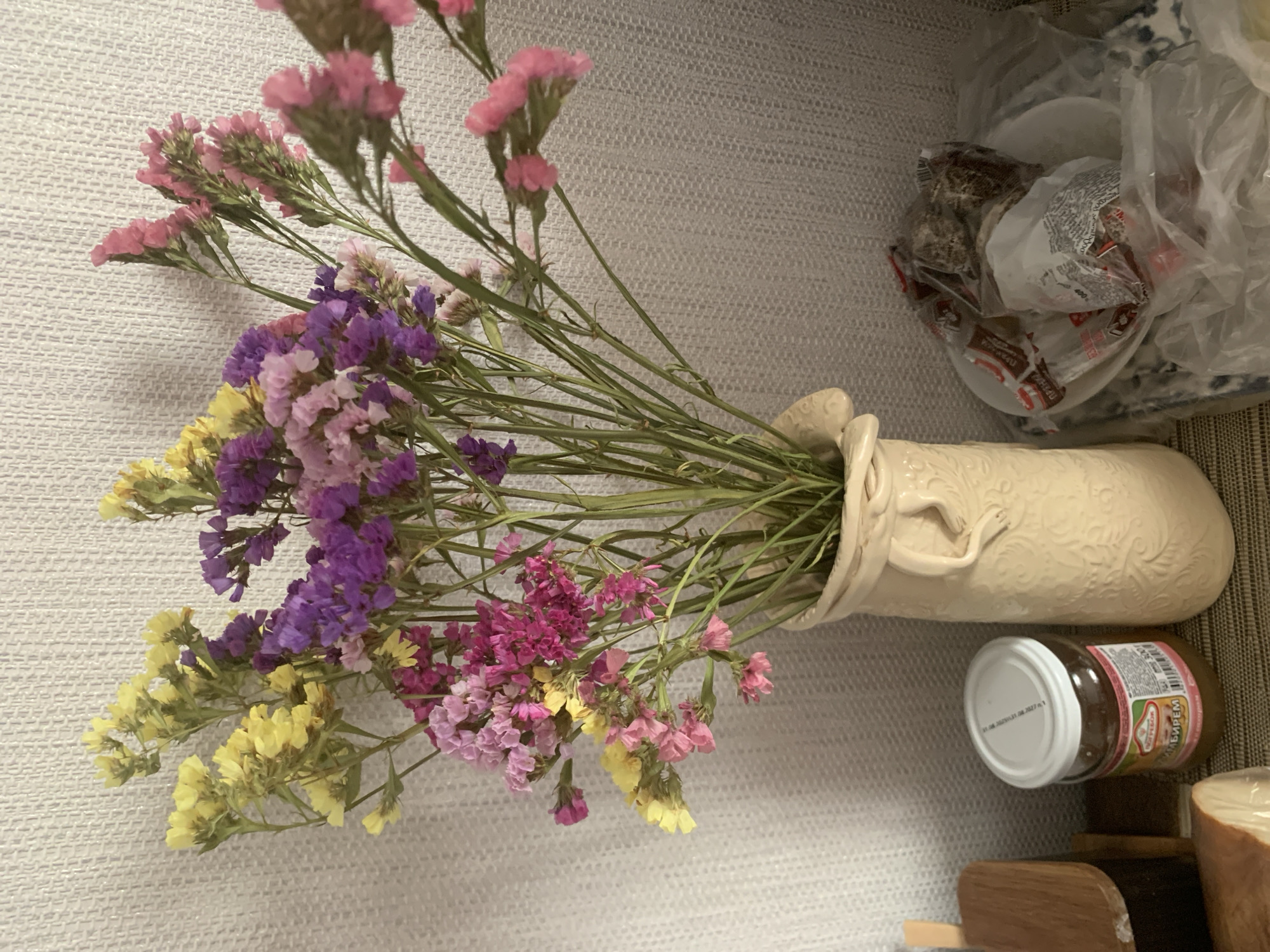 A still life composition features a cream-colored vase filled with dried flowers in shades of yellow, purple, and pink. The vase has an embossed pattern and is placed on a surface next to a small glass jar with a white lid, which appears to contain jam. To the right of the vase, a white plate holds several items wrapped in plastic packaging. In the background, a textured wall provides a neutral backdrop. The image is taken indoors, likely in a residential setting.