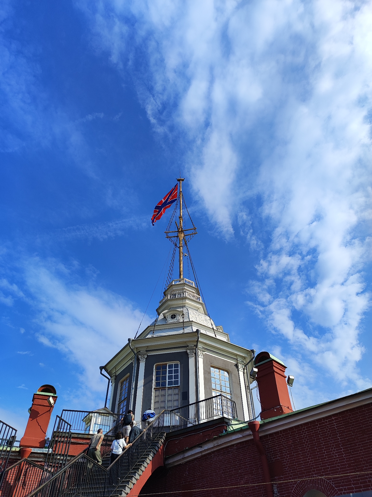 A Dutch flag flies from a mast atop a building in Saint Petersburg, Russia, with a bright blue sky and scattered white clouds overhead. The building, with its grey facade, white trim, and multi-paned windows, appears to be part of a historical complex, possibly a fortress or naval facility, given the flag's design and the mast. Red brick walls and a red chimney structure with a green roof are visible in the foreground. A set of metal stairs with black railings leads up to the building, and several people are ascending them. The perspective is from a low angle, looking up at the flag and the building.
