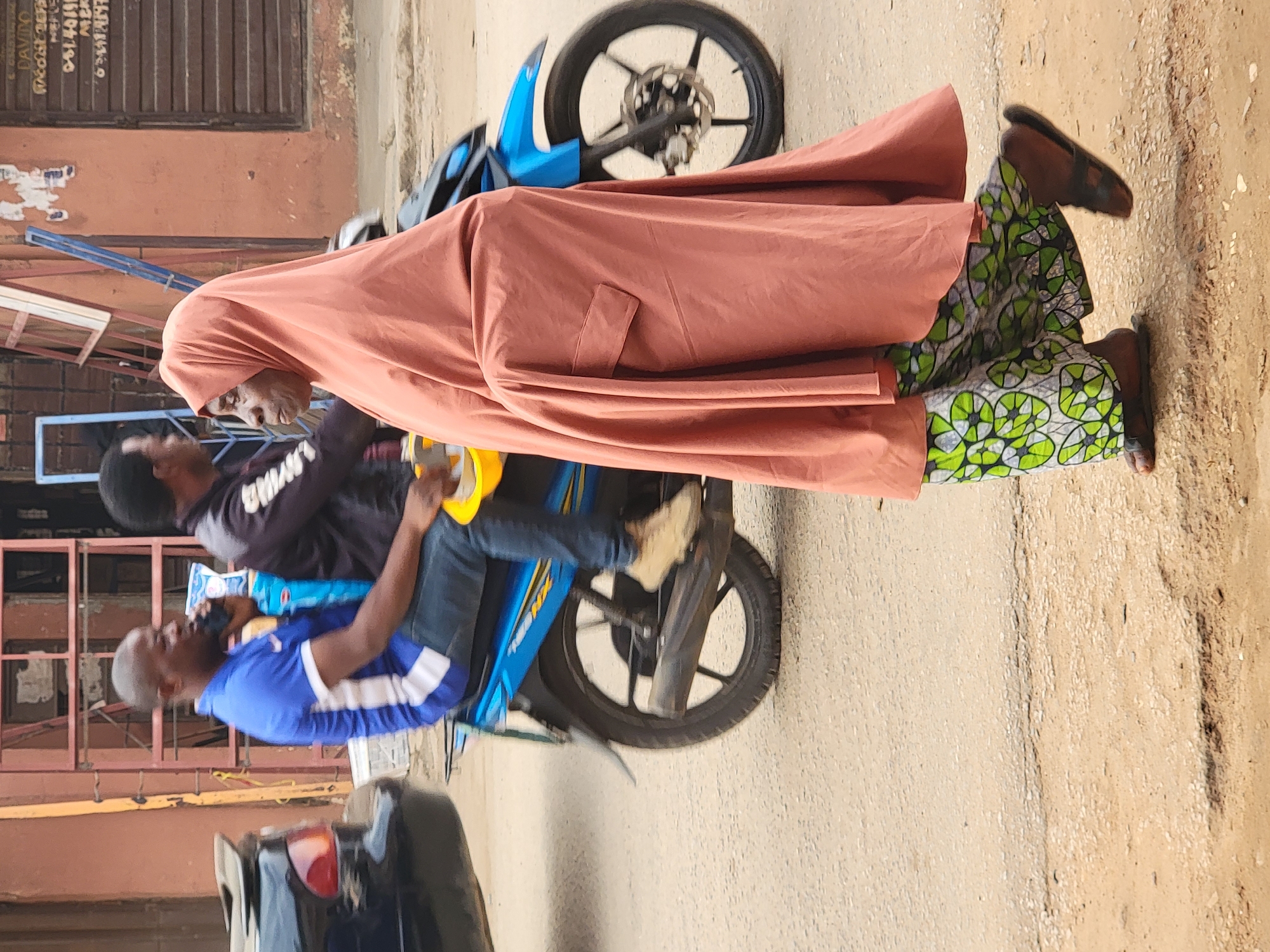The image captures a vibrant street scene in Maiburiji, Nigeria, under natural daylight. In the foreground, a woman stands on the right, adorned in a long, flowing salmon-colored robe that covers most of her body, revealing a striking green and white patterned skirt or dress underneath. She wears dark sandals, and her partially visible face suggests an elderly individual with a calm expression.
Beside her, two men are seated on a blue "TVS NTORQ 125XL" motorcycle. The driver, with short grey hair, is wearing a blue t-shirt with white stripes and blue jeans. He holds a yellow roll, possibly tape or another utility item, in his right hand. The passenger behind him is dressed in a dark long-sleeved shirt with white lettering.
The setting is an urban or semi-urban street, with a mix of concrete and dirt ground. In the background, buildings with shuttered windows and doors are visible, displaying signs of age. Visible text on one of these buildings includes "DAVIDO," followed by numbers and words like "0814011," "MERCH," "O.C.A.S.," and "ENGRS." A portion of a black car is visible in the lower-left corner. The overall scene depicts a casual moment of daily life or a brief stop on a local thoroughfare.