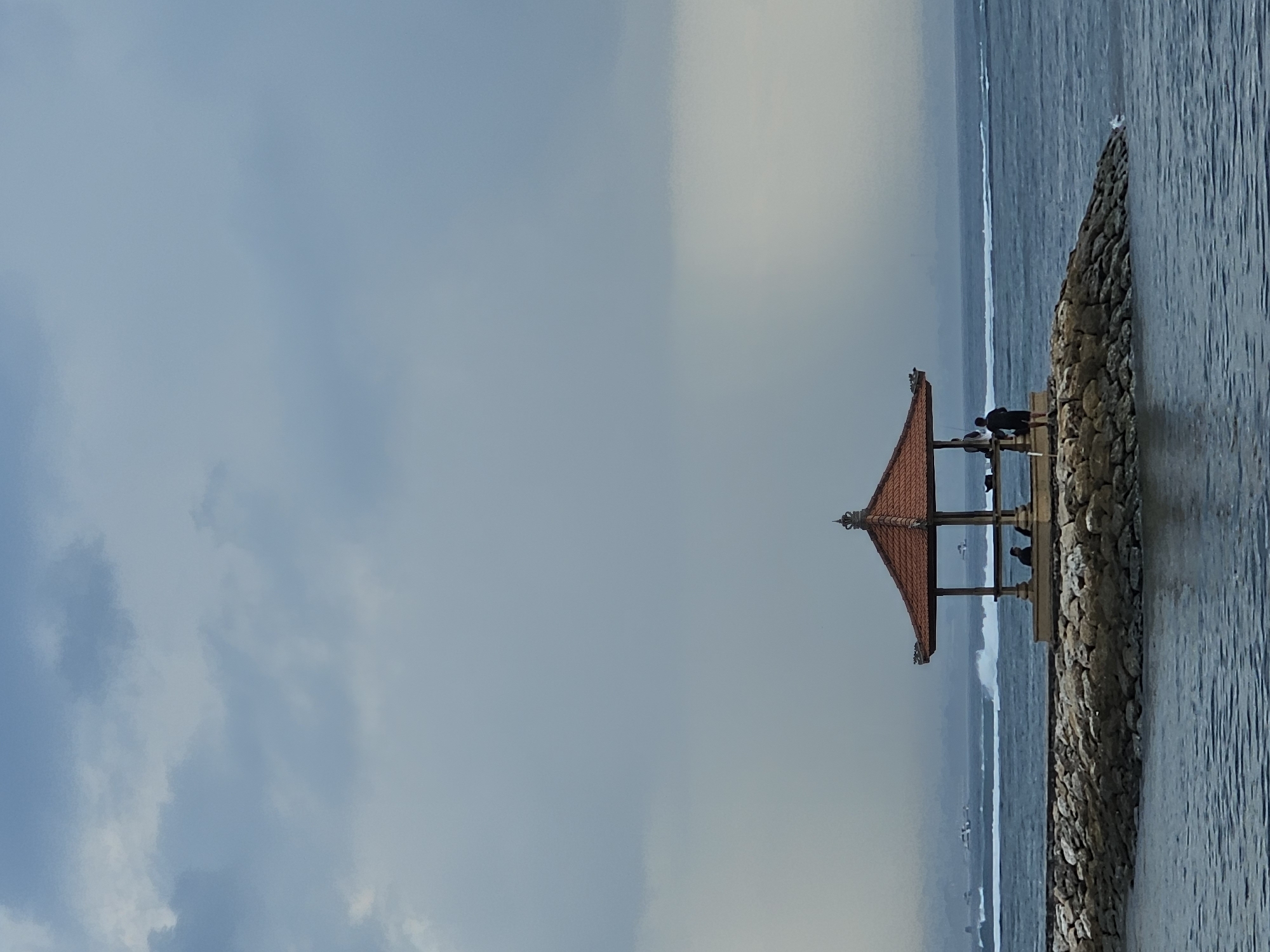 The image captures a serene scene at the Grand Welingan Temple area in Denpasar, Indonesia, likely during a cloudy day. In the foreground, the dark blue water of the ocean laps against a rocky shore. Prominently situated on this rocky outcrop is a traditional Balinese pavilion with a tiered, red-tiled roof. The pavilion has an open design with visible wooden supports and decorative elements at the apex of the roof.
Within the pavilion, two figures are visible. One person appears to be seated, while another stands. Outside the pavilion, to the right, a third individual stands, facing away from the camera, seemingly observing the sea.
In the mid-ground, the ocean stretches towards the horizon, with several boats and their wakes visible in the distance, suggesting moderate activity on the water. The sky above is filled with heavy, gray clouds, creating a muted and atmospheric light. The overall impression is one of tranquility and natural beauty, juxtaposed with the man-made structure and the distant signs of human presence.