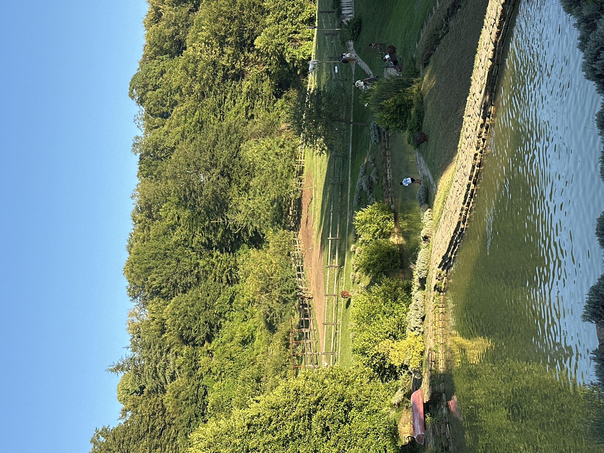The image depicts a serene countryside scene, possibly a farm or estate, with a clear blue sky overhead. In the foreground, a tranquil pond reflects the surrounding greenery and a stone path winds across its surface. To the left of the pond, a red boat rests on the grassy bank, near a cluster of lush plants.
The middle ground features a gently sloping lawn dotted with bushes and trees. A wooden fence encloses a pasture where a white horse grazes. A group of people are seen walking along another stone path that leads up the incline, some pausing to admire the scenery or rest on a bench.
The background is dominated by a dense forest of mature trees, their vibrant green foliage creating a natural backdrop. The overall impression is one of peace and natural beauty, with a hint of human activity integrated into the landscape. The location is identified as the city of Progress, Russia.