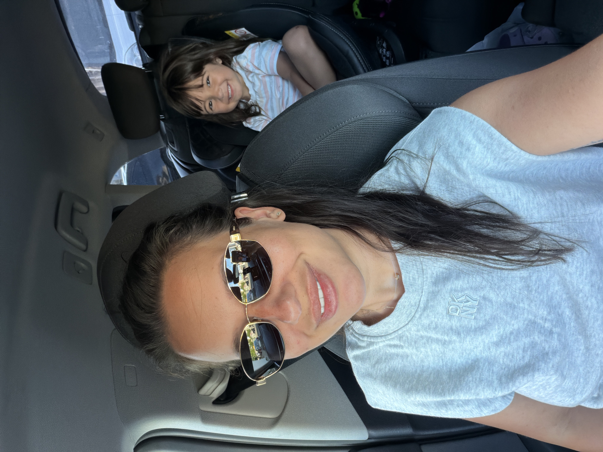 In the front passenger seat of a car, a woman wearing sunglasses and a light grey t-shirt is taking a selfie. She has dark hair pulled back and is smiling. In the back seat, a young girl with brown hair is seated in a car seat, also smiling at the camera. The interior of the car appears to be a modern vehicle, with dark grey upholstery and visible headrests. Reflections in the woman's sunglasses show a street scene with cars and buildings, suggesting the car is in motion or parked on a street. The setting of the image is within the city of Burgas, Bulgaria, based on the provided metadata.