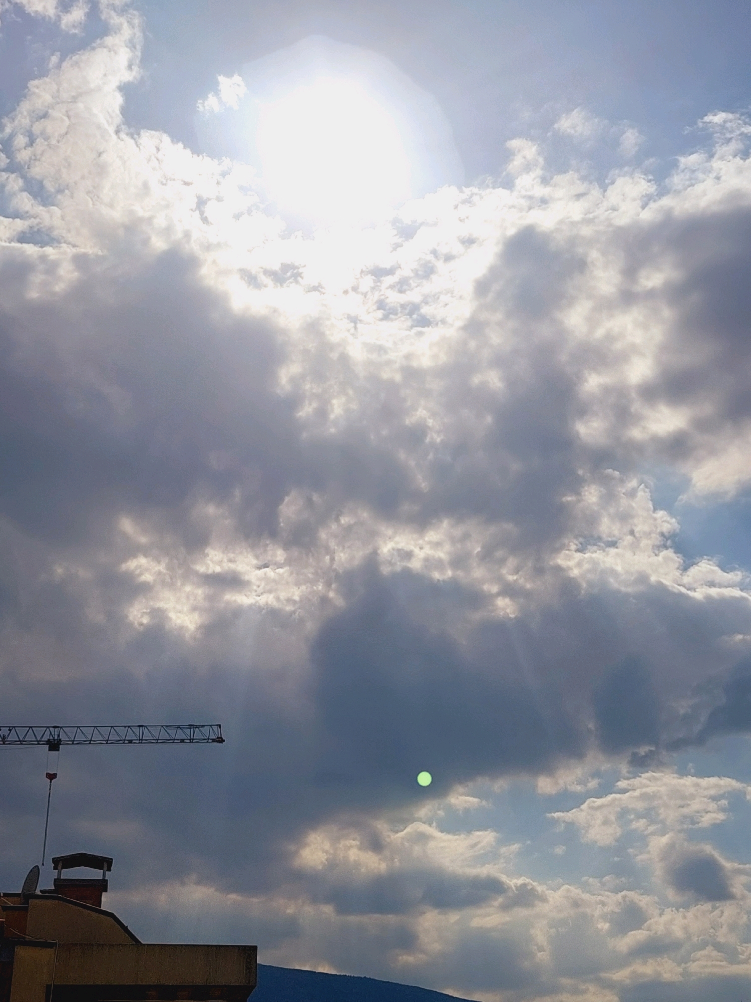 The image captures a cloudy sky with the sun partially obscured, creating a diffused light. Ray of light are visible, beaming through the clouds. In the foreground, the top of a building with a chimney and a satellite dish is visible. To the left, a construction crane extends horizontally across the frame. A small, bright green circular object is present in the sky. The scene is likely a city landscape, possibly Sofia, Bulgaria, given the context provided.