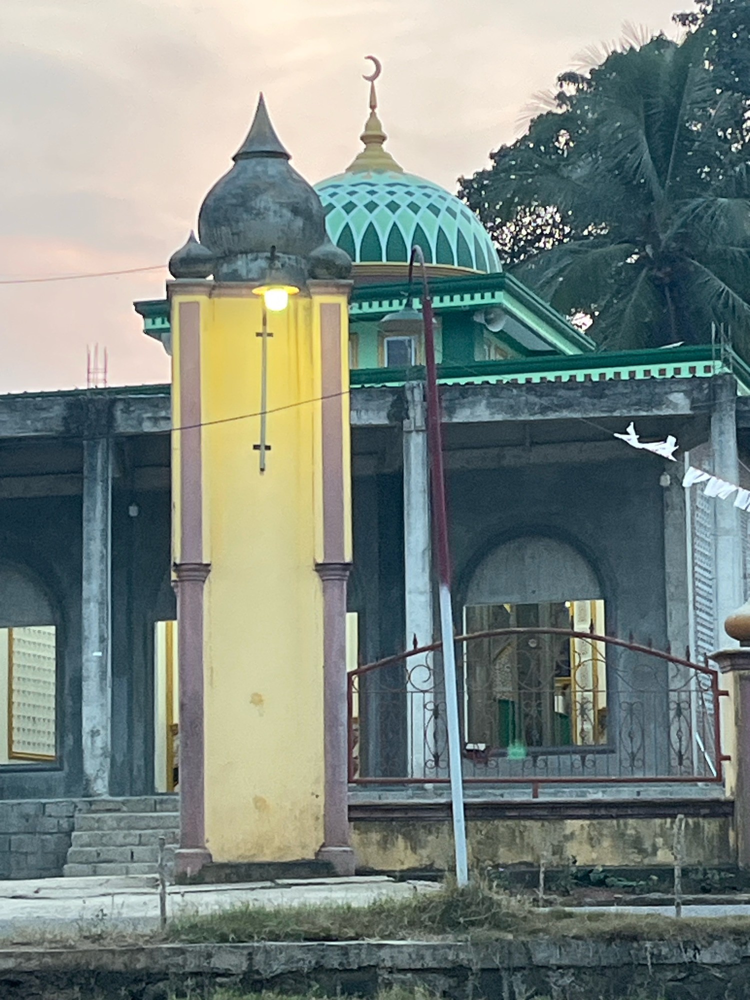 The image captures the exterior of a mosque, possibly located in Kebumen, Indonesia, during twilight. A tall, slender minaret-like structure painted in yellow with maroon vertical accents stands prominently in the foreground to the left. It is topped with a dark, conical dome. A light fixture is attached to the side of this structure. Behind it, the main mosque building is visible, featuring a green dome adorned with diamond-shaped patterns and a crescent moon finial. The building itself has a green roofline and a grey, unfinished concrete facade with arched windows. A red metal fence with decorative swirls is positioned in front of these windows. A tall, thin pole with red and white markings stands vertically in front of the building and fence. In the background, lush green trees, including a palm tree, are visible against a pale, overcast sky. The ground in front of the mosque appears to be paved and there is a body of water or a canal in the immediate foreground. A piece of white paper, possibly a decoration or kite in the shape of an airplane, is attached to a string and visible to the right of the mosque's main structure. Small white flags are also strung along the building.